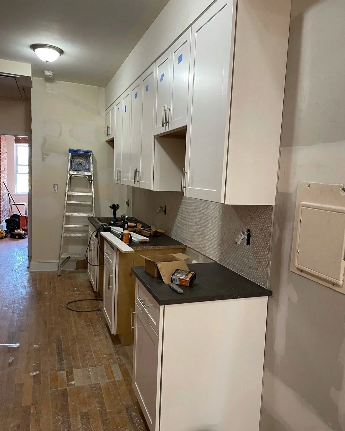 Kitchen under renovation with white cabinets, tools, and materials on the countertops, a ladder in the background, and ongoing work.