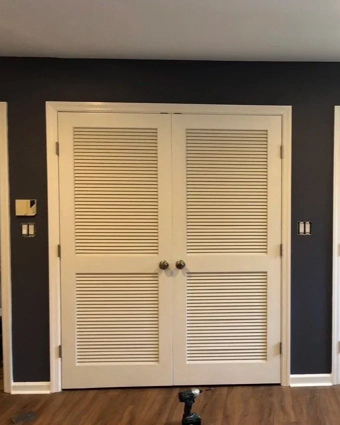 Closed white louvered double doors in a room with dark navy walls and hardwood flooring.