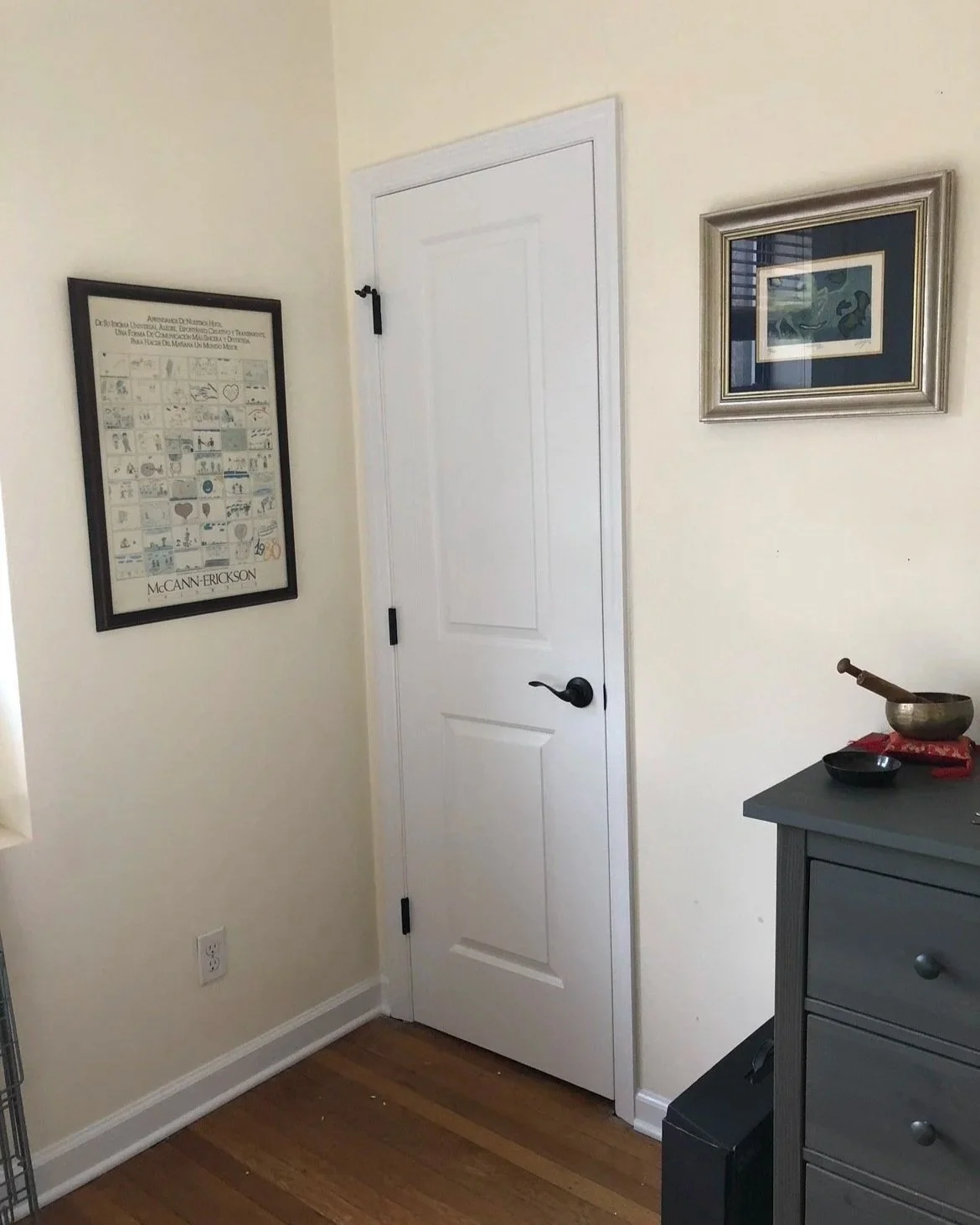 A room with a white door, framed artwork on the walls, and a gray dresser with a singing bowl and small black dish on top.