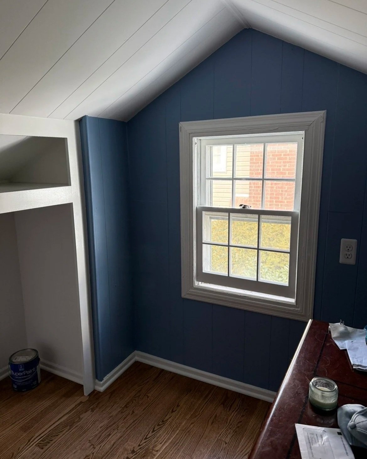 Corner of a room with blue walls, white window frame, and a small section of a wooden floor. A window shows an outside view of a brick building. Part of a desk or table with papers and a candle is visible.