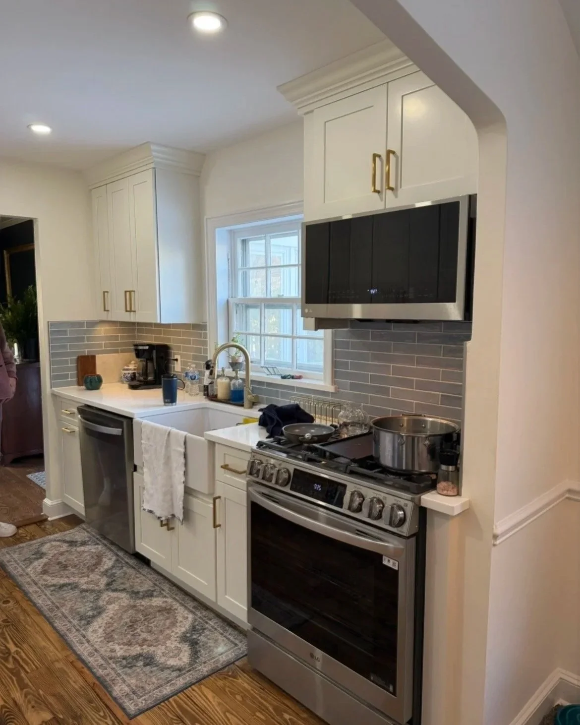 A kitchen with white cabinets and a gray brick tile backsplash, a window over the sink, a stainless steel oven and microwave, and various small appliances and items on the counter.
