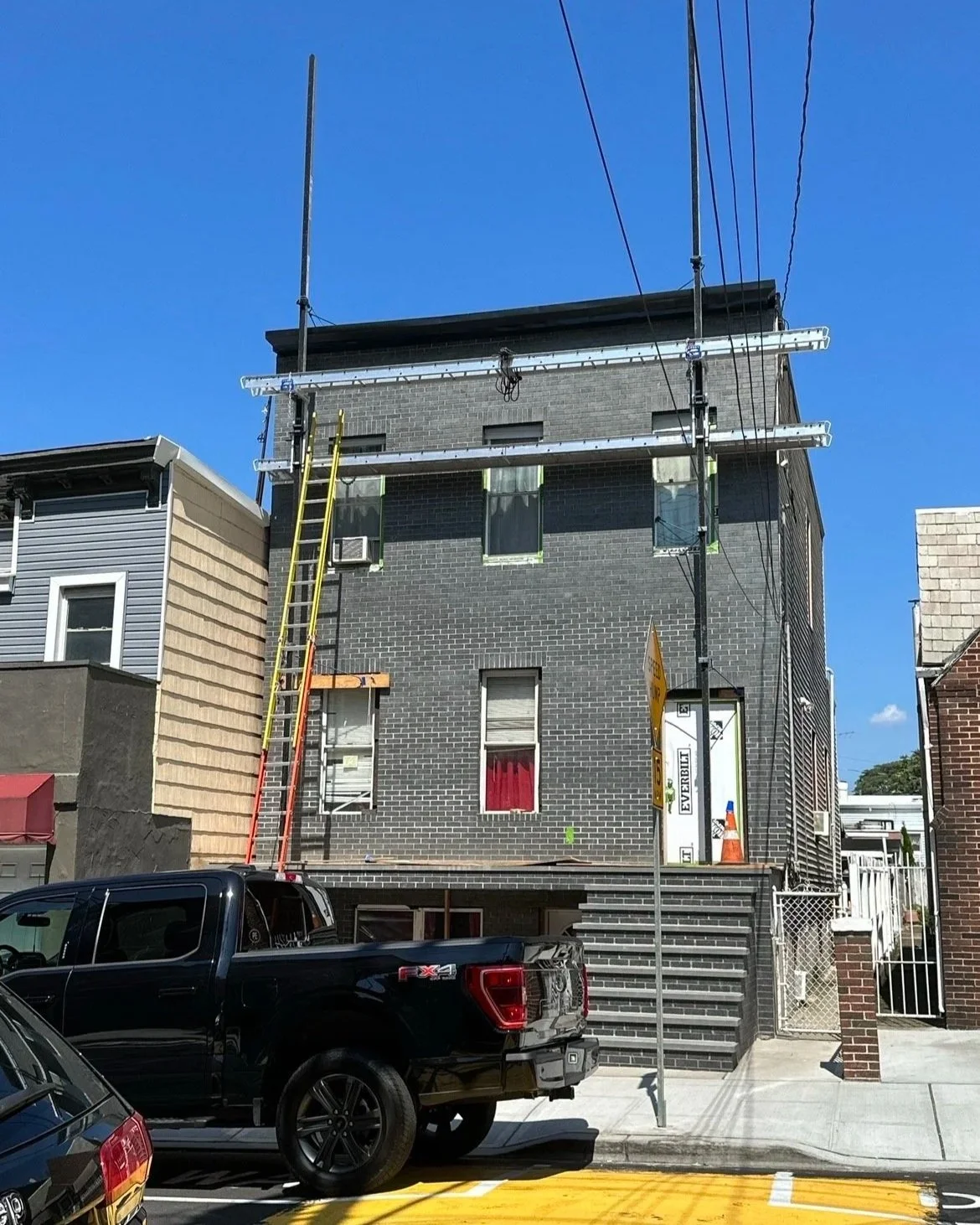 Building under construction with scaffolding and ladders, parked cars in front, and a signboard on the sidewalk.