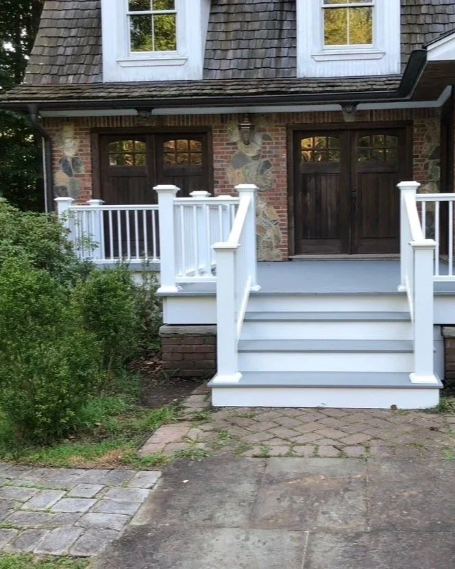 Front view of a house with a staircase leading to a porch, white railings, brick steps, and dark wooden door and window shutters.