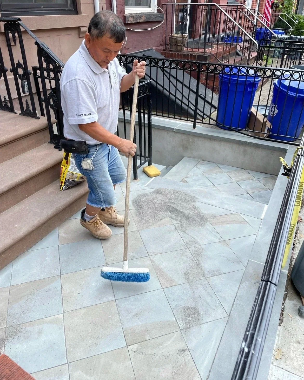 A man cleaning the front porch of a house with a broom, wearing a white shirt, blue jeans shorts, and tan work boots.