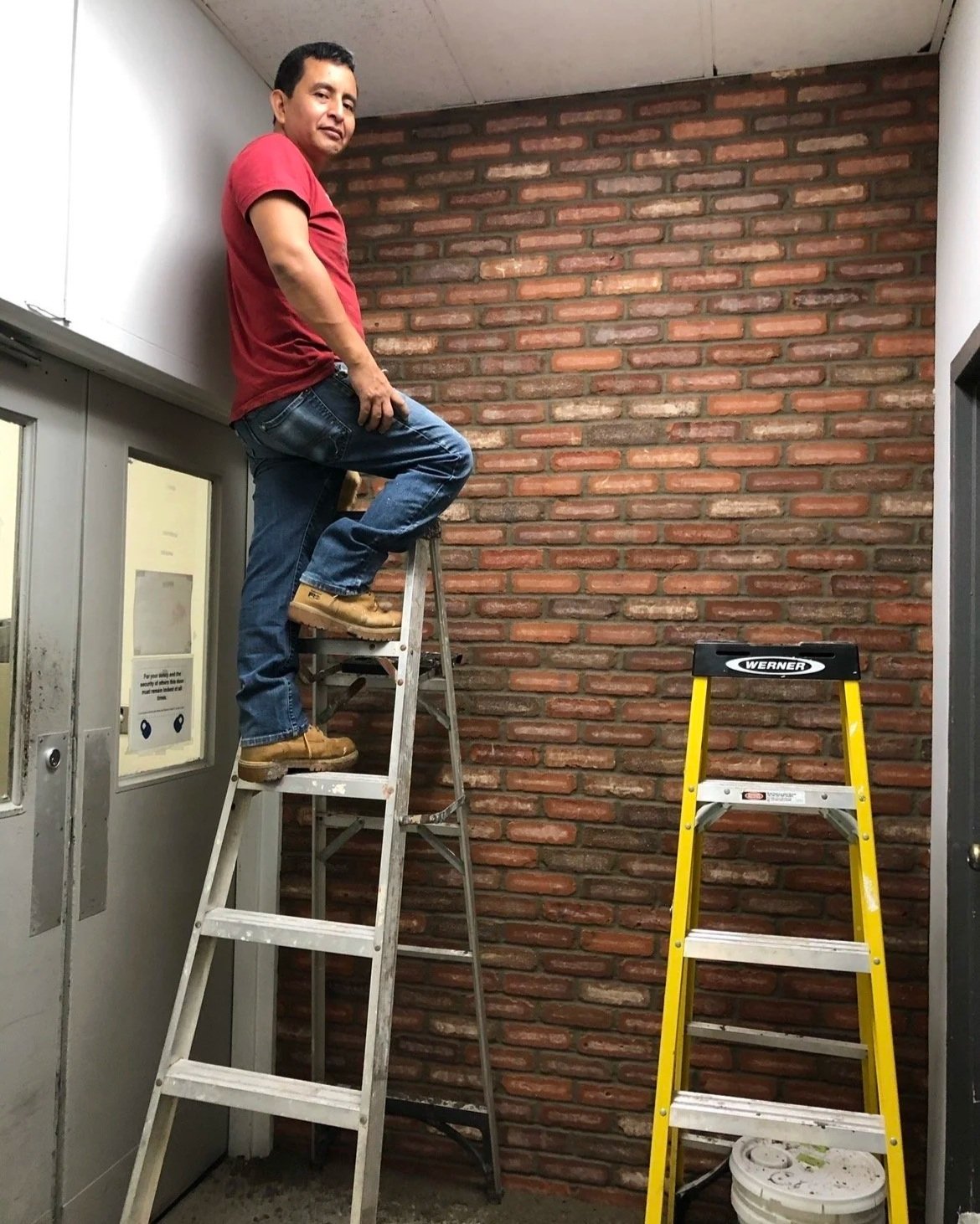 A man standing on a step ladder in front of a brick wall, with another empty yellow step ladder nearby.