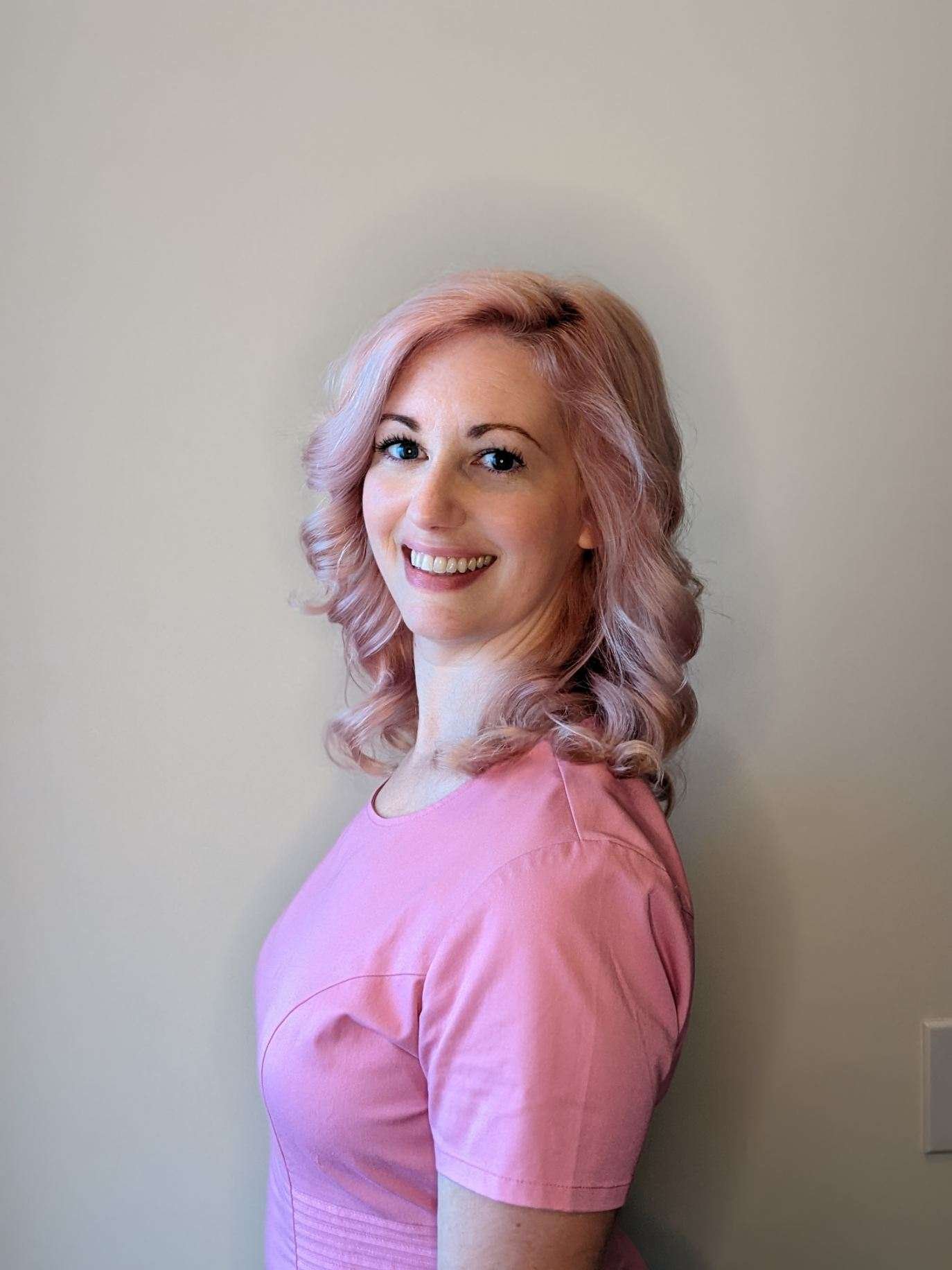 Person with pink hair wearing a pink shirt, smiling in side profile against a plain background.