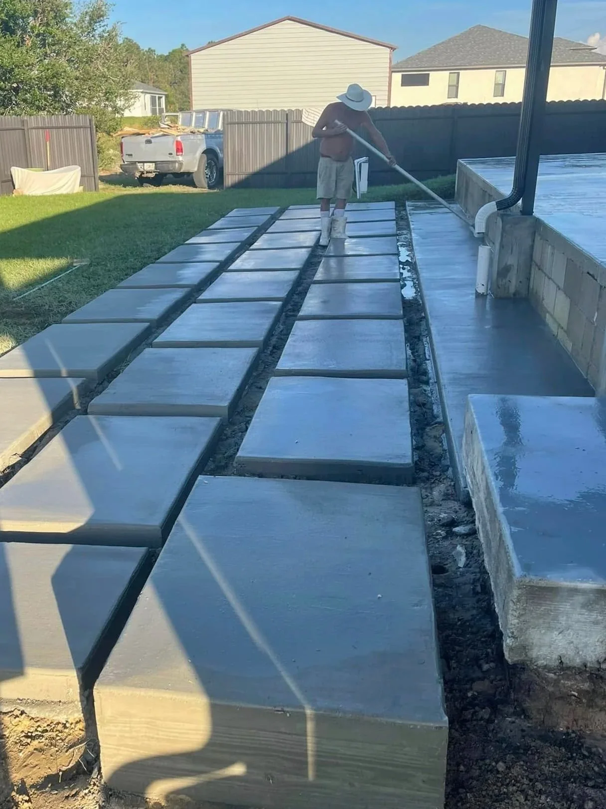 Person laying down concrete pavers for a new patio or walkway, with a worker spreading concrete with a long tool.