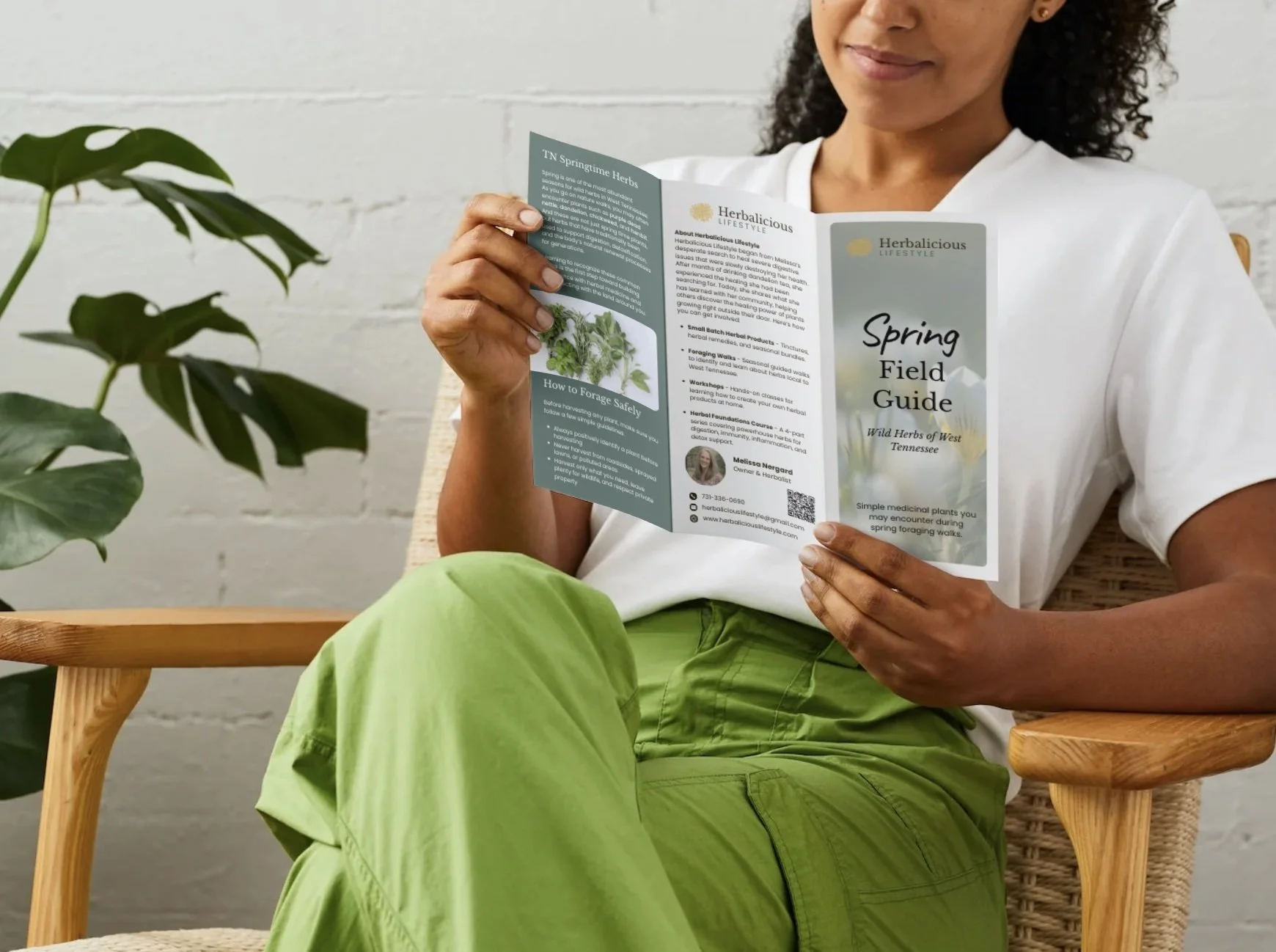 Woman sitting on a wooden chair, holding a spring field guide booklet with herbal information, next to a green plant, with a white painted brick wall background.