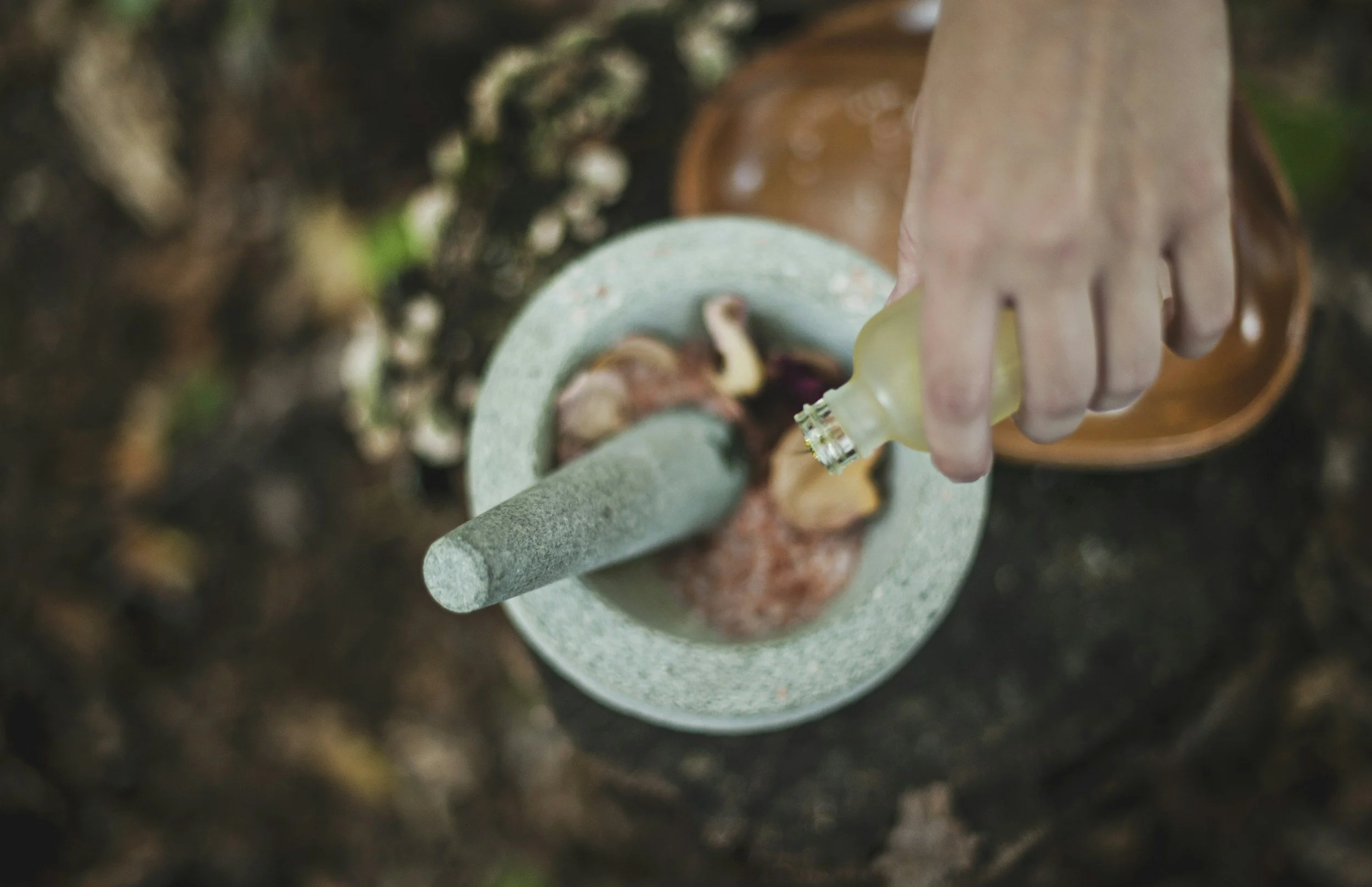 A person using a mortar and pestle to grind herbs or spices with a hand holding a dropper bottle nearby.