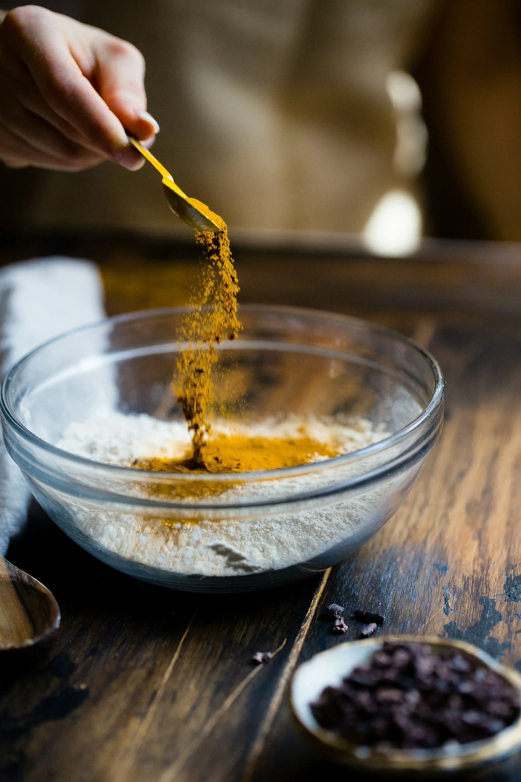 A person adding yellow turmeric powder to a glass bowl containing white flour on a wooden table.