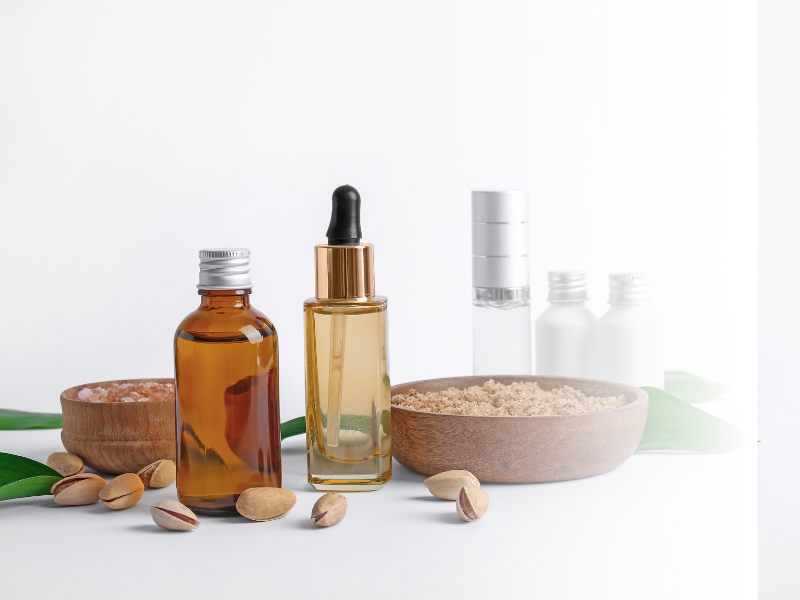 Assorted natural skincare products including a dropper bottle, a small amber bottle, and two white bottles, with salts and green leaves on a white surface.