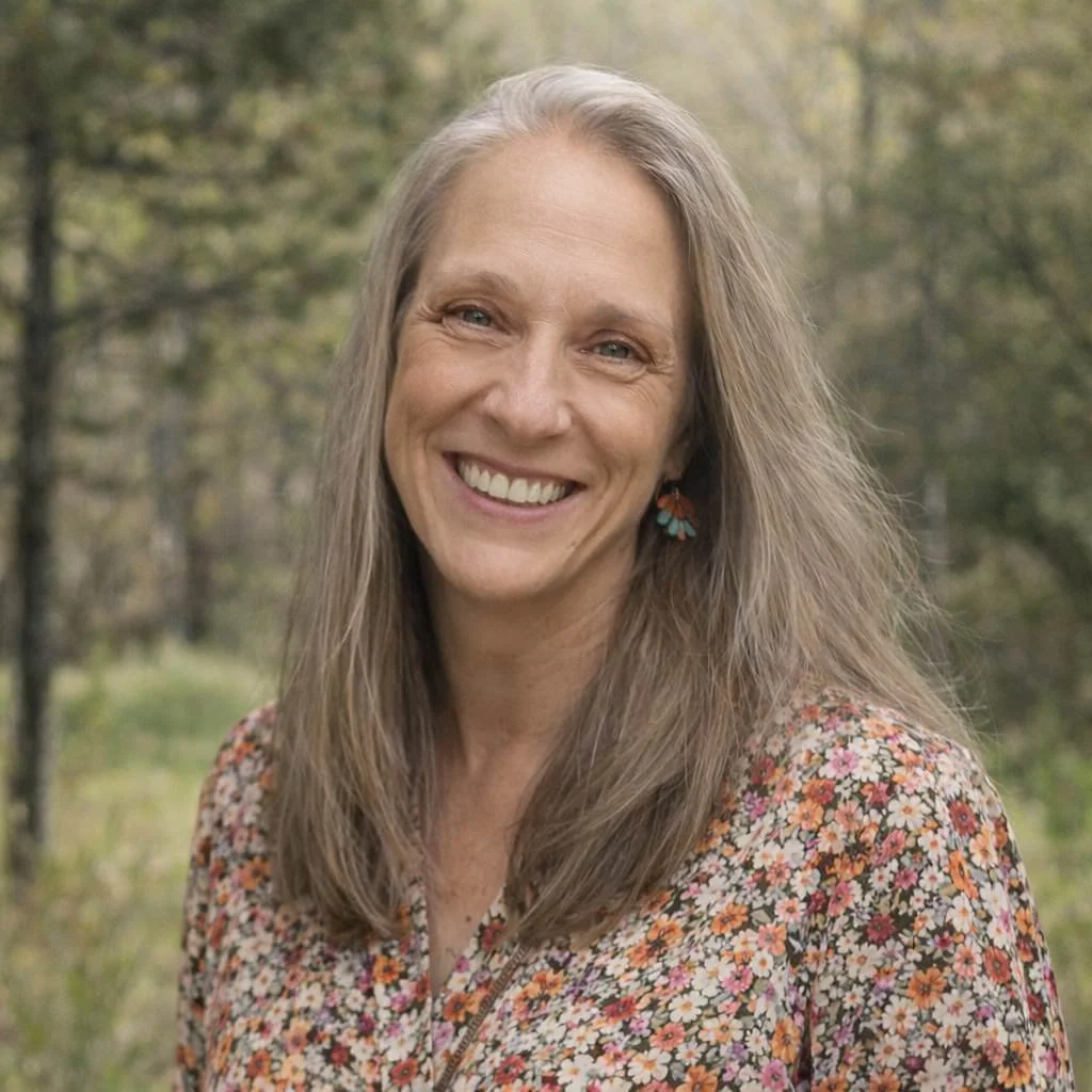 A smiling woman with long gray hair wearing a floral blouse and colorful earrings in an outdoor setting with trees in the background.