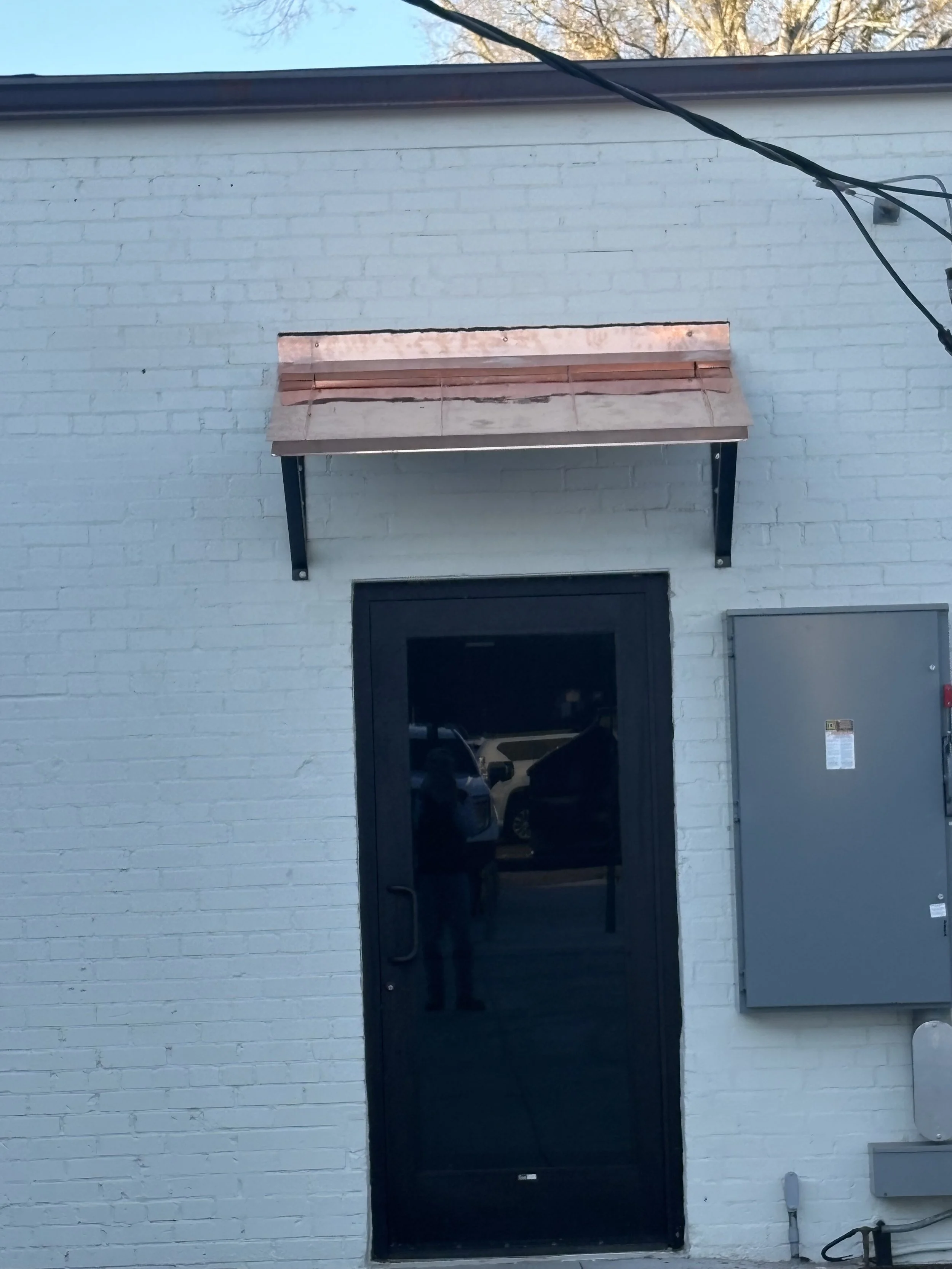 Black door on a white brick wall with a small copper-colored awning above it, a gray electrical box on the right, and power lines overhead.