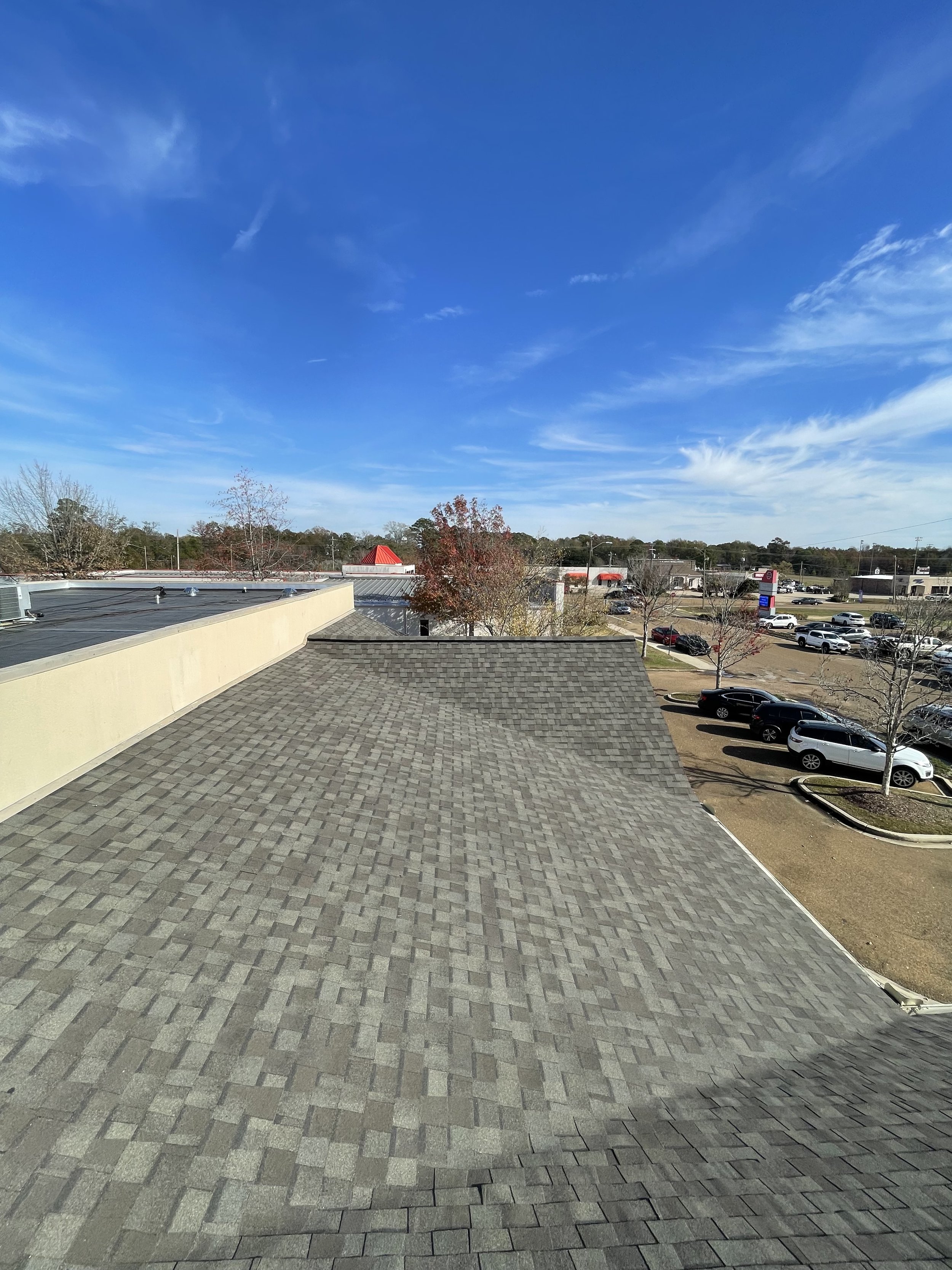 View of a parking lot with cars and trees, under a blue sky with some clouds of a building's roof in the foreground.