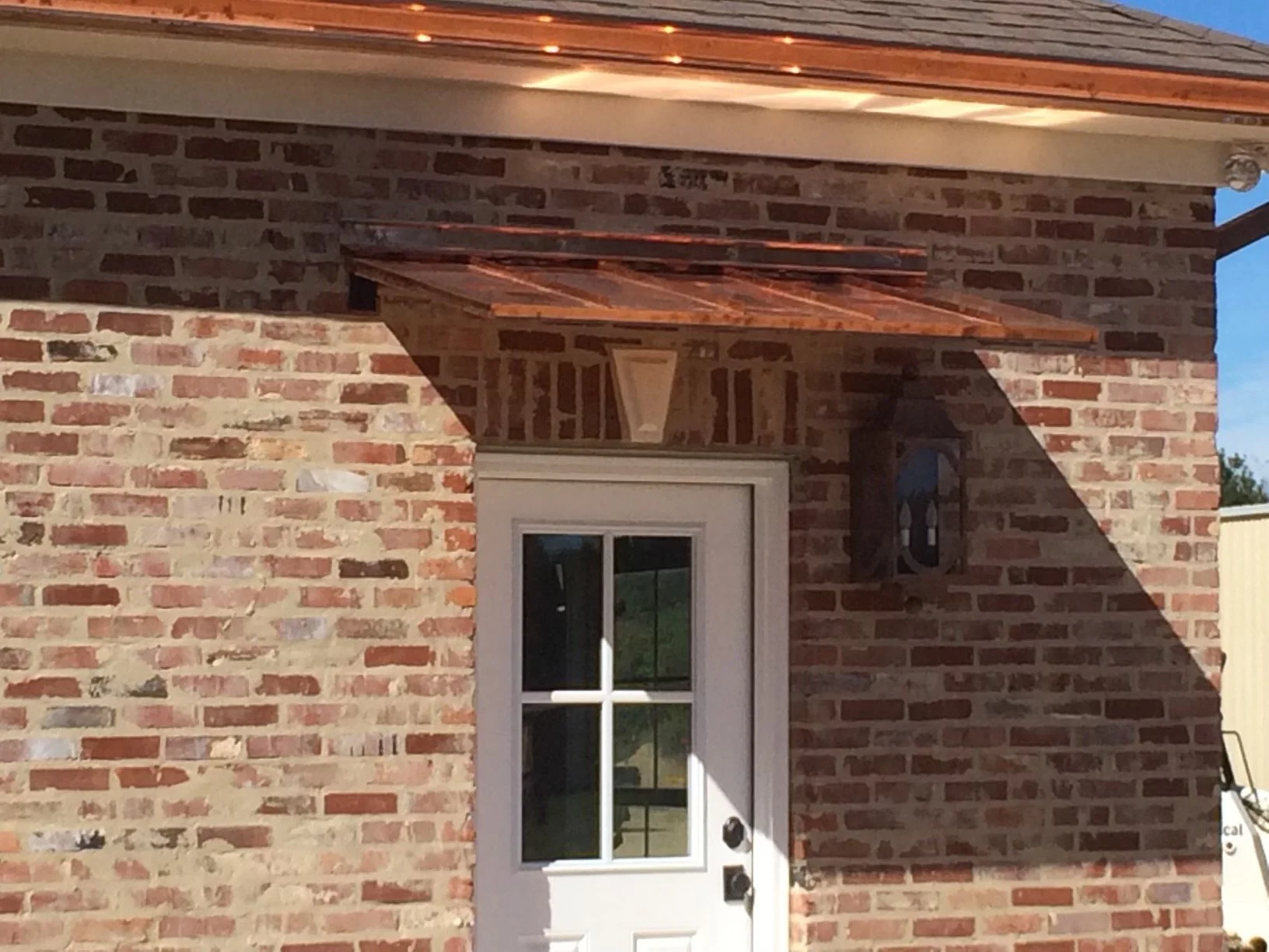 Close-up of a brick house exterior with a white door, a black lantern-style light fixture, and a small copper awning above the door, casting a shadow on the wall.
