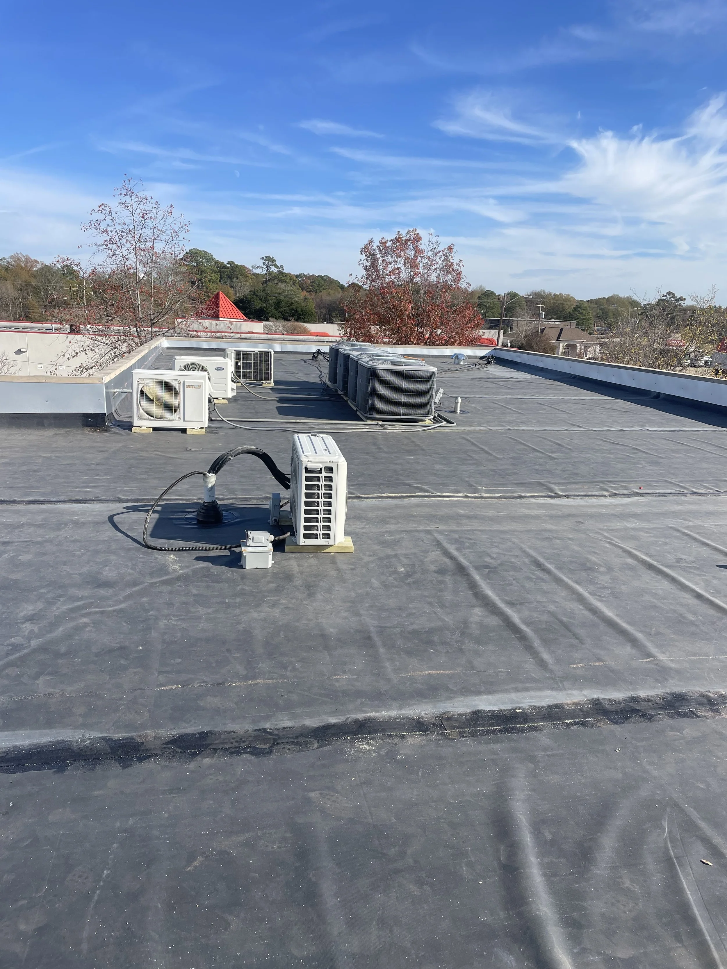 Flat rooftop with multiple HVAC units and air vents, under a clear blue sky with trees and rooftops in the background.