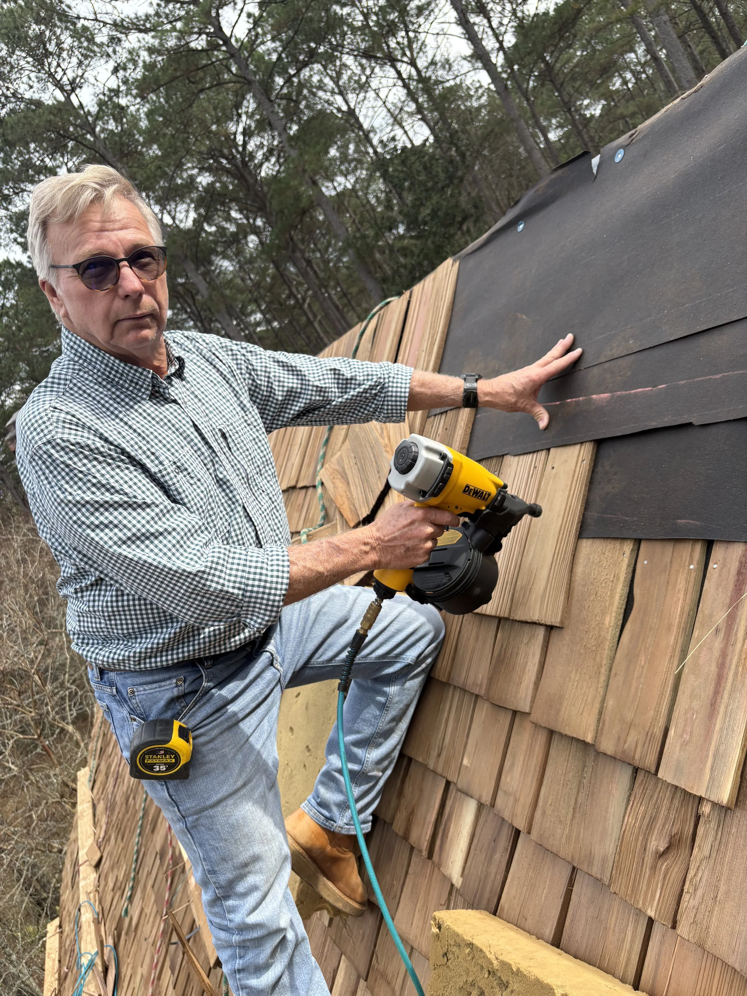 A man in sunglasses installing or repairing shingles on a roof using a power nail gun, with roofing materials and trees in the background.
