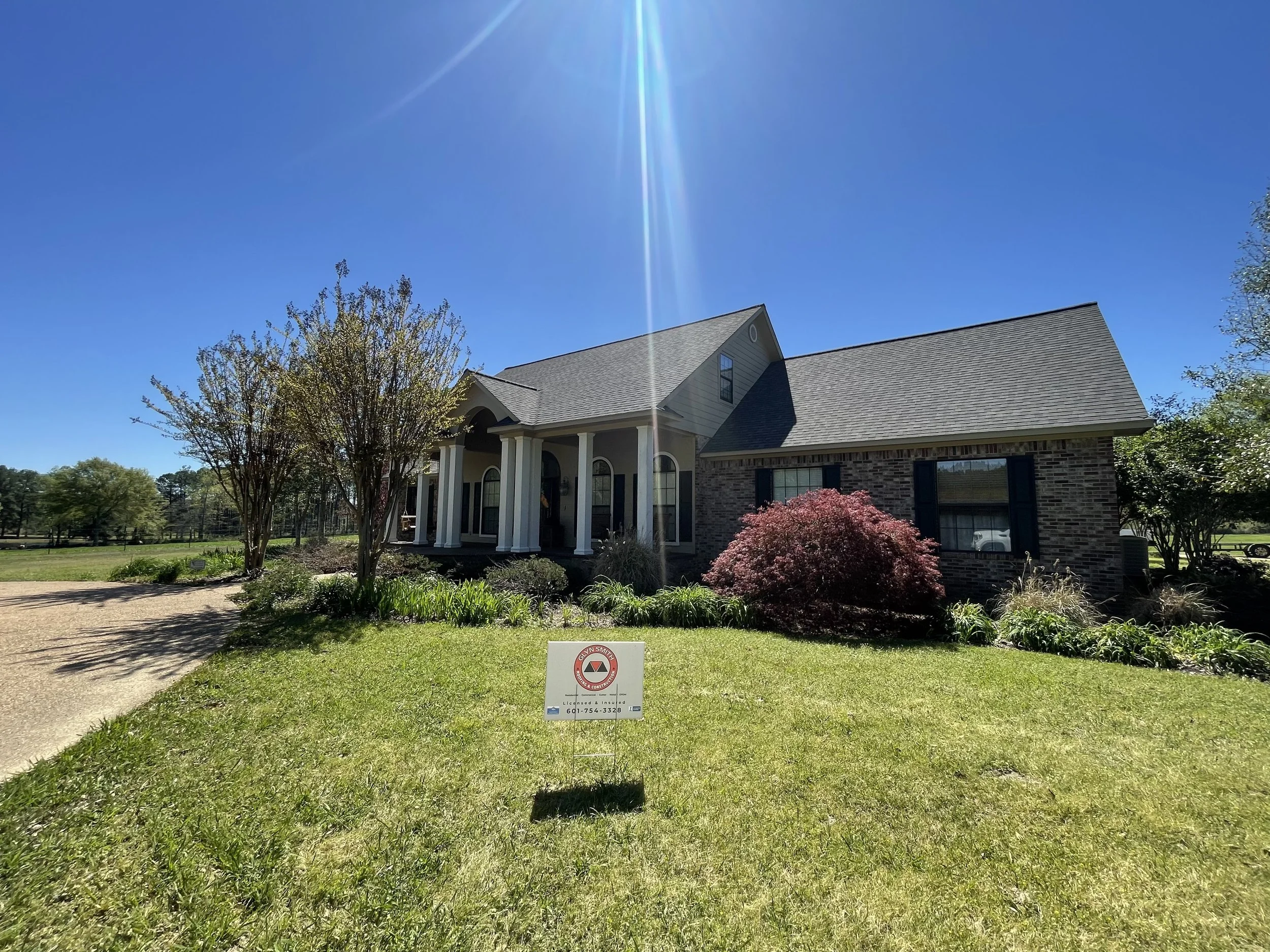 A house with brick and siding exterior, front porch, and landscaped yard under a clear blue sky with the sun shining.