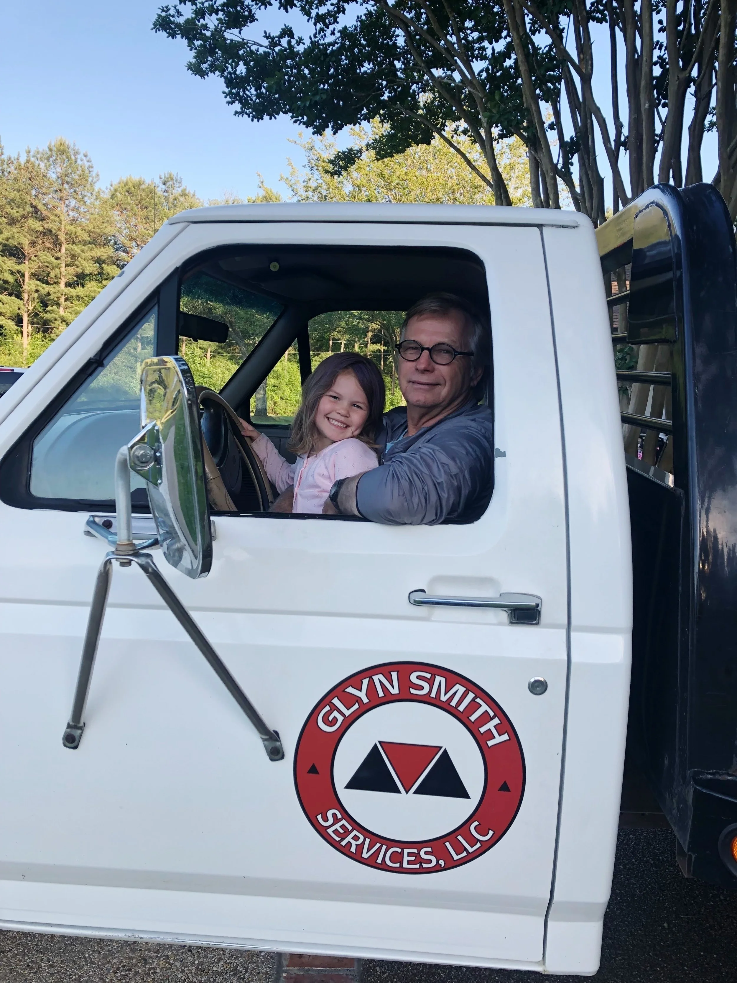 A young girl and an older man are sitting in the cab of a white service truck with a logo that says Glyn Smith Services, LLC. They are smiling, and the girl is holding the steering wheel. The scene is outdoors with trees and a blue sky in the backgro