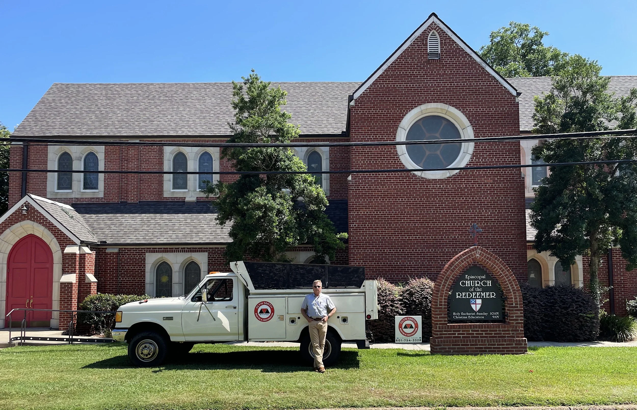 A man standing next to a white utility truck in front of a red brick church building with a large circular window and a sign that reads 'Episcopal Church of the Redeemer' under a cross, with trees and a bright blue sky in the background.