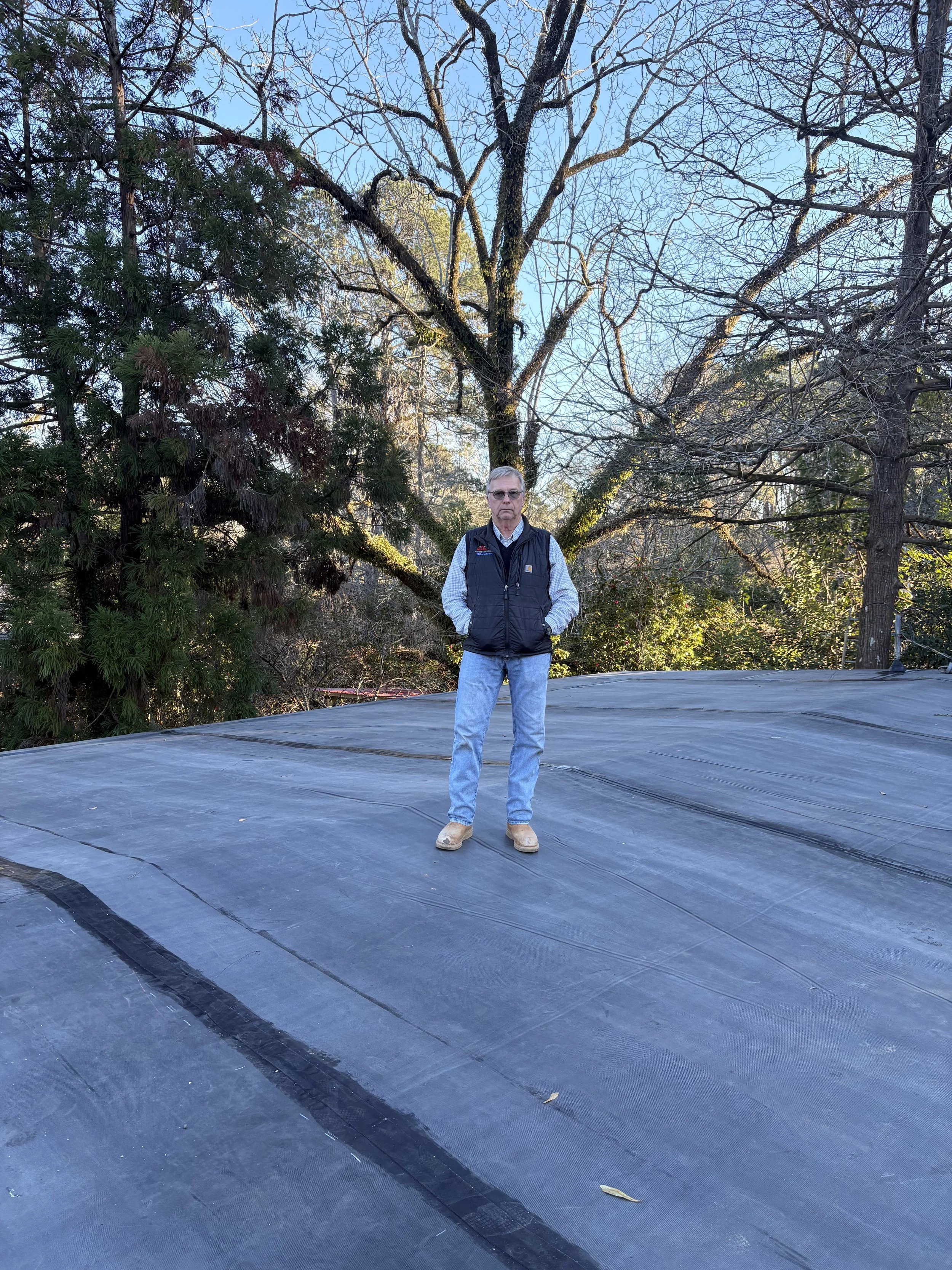A man standing on a flat, black roofing material outdoors with trees and a clear blue sky in the background.