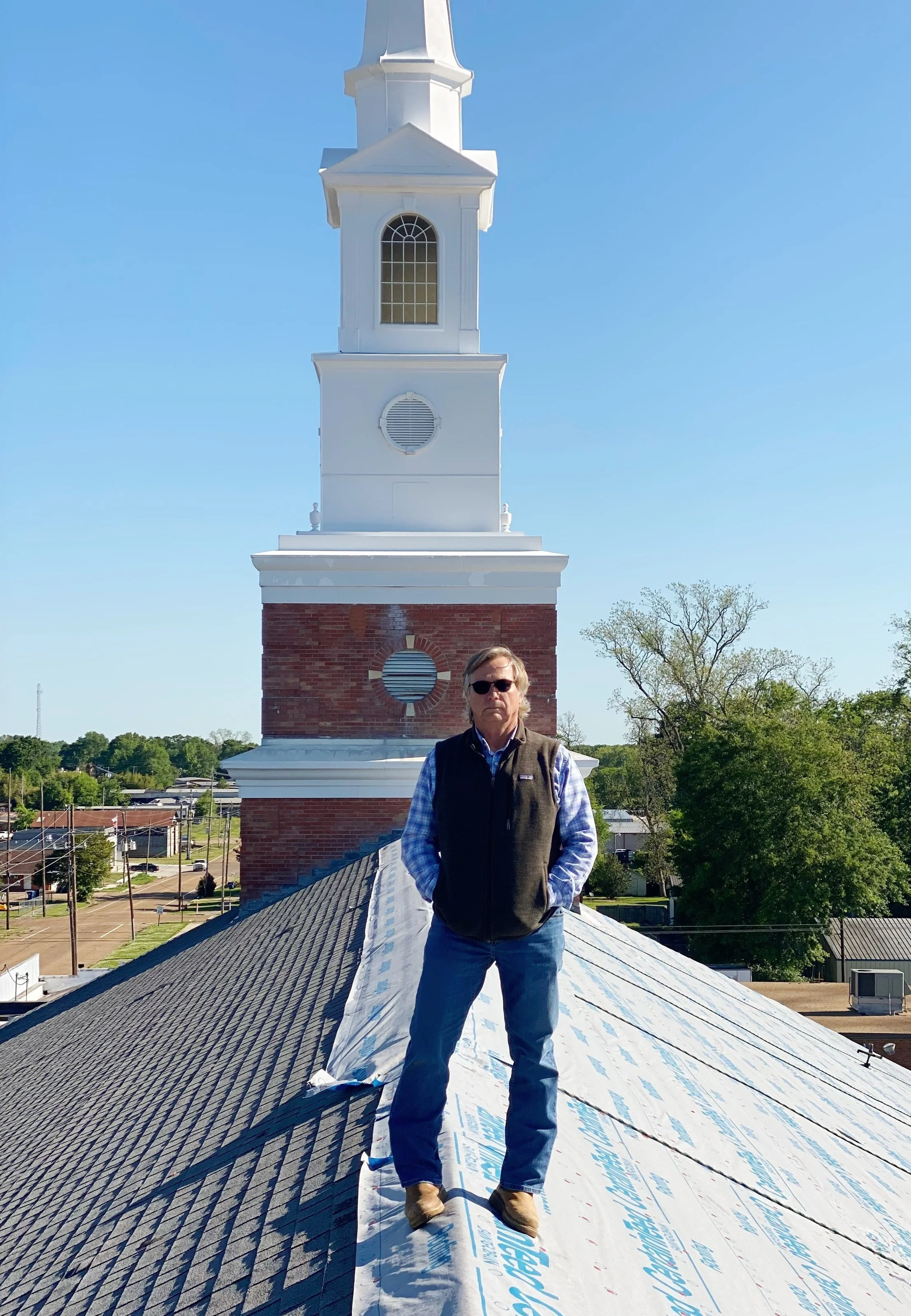 A man stands on a roof under construction, wearing sunglasses, a black vest, a blue plaid shirt, jeans, and boots, with a church steeple and blue sky in the background.