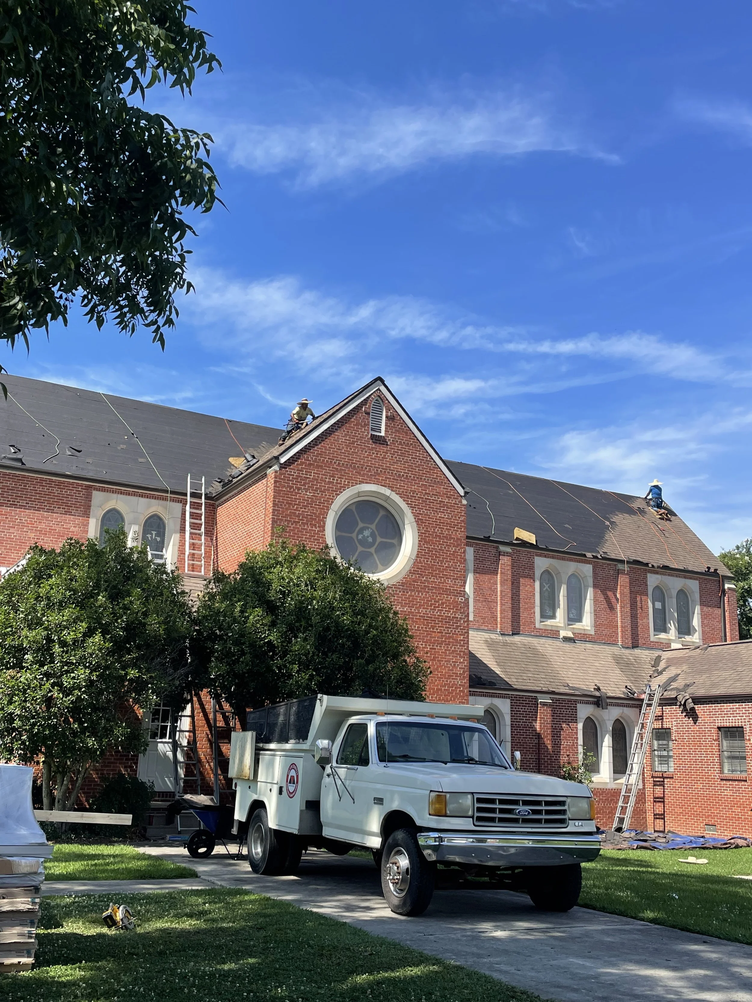 Workers repairing the roof of a red brick church on a sunny day with blue sky, a white utility truck parked in front, ladders leaning against the building, and trees nearby.
