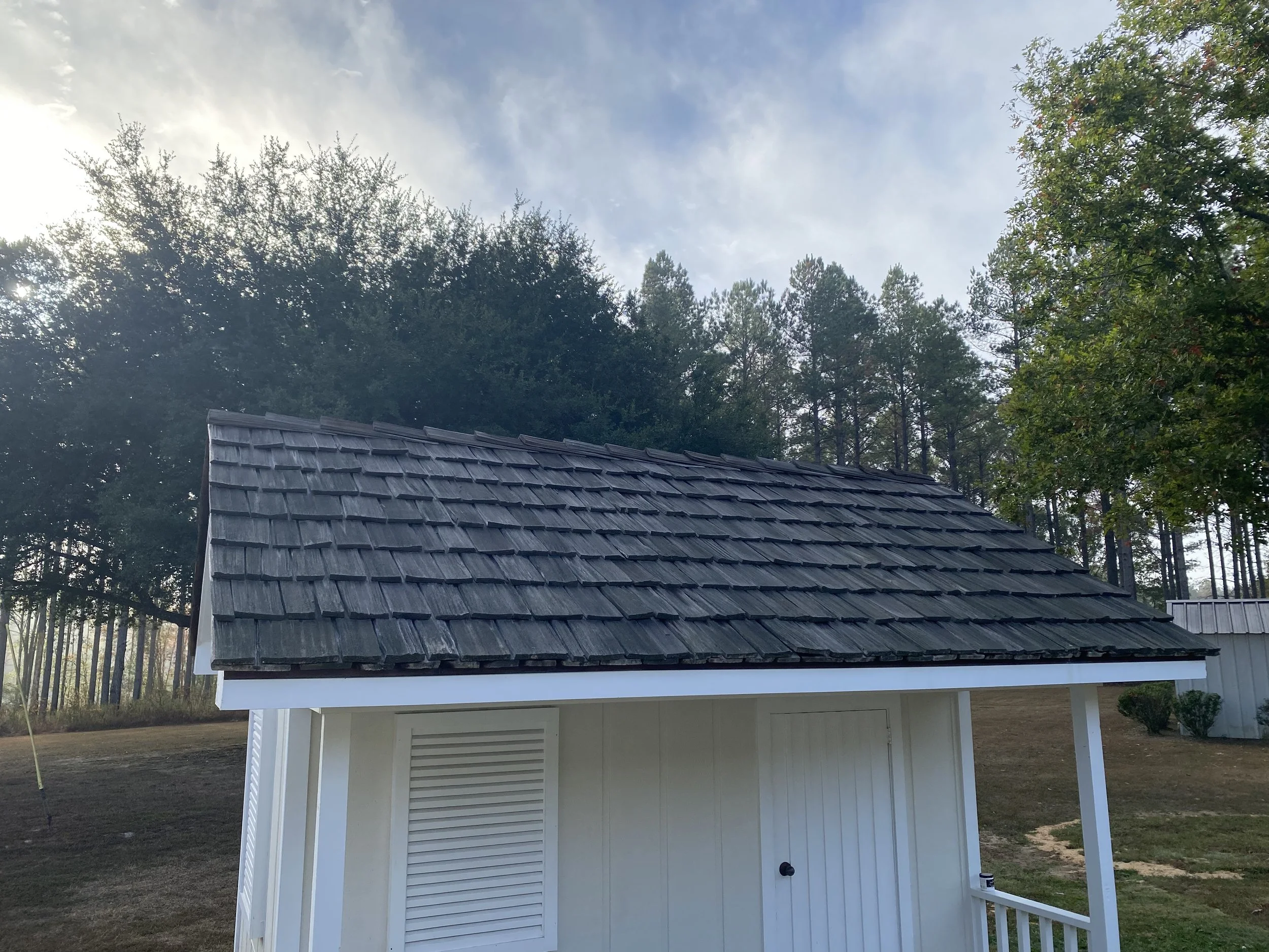 Small white building with a dark gray shingle roof, located outdoors with trees in the background and a partly cloudy sky.