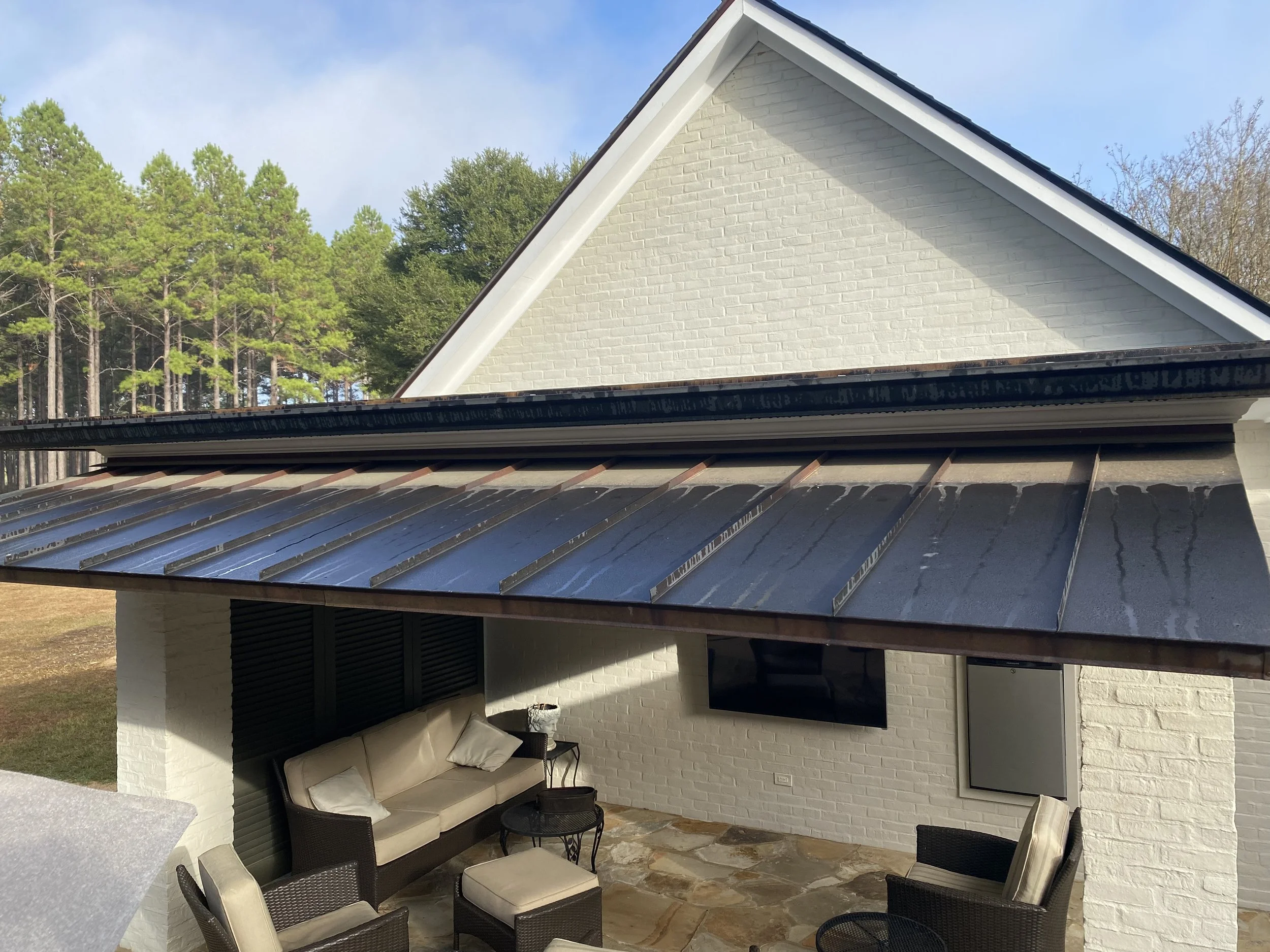 Outdoor patio area with seating, including a sofa and chairs, under a roof with a metal ridge cap, attached to a white brick house, with a large TV on the wall and a forested background.