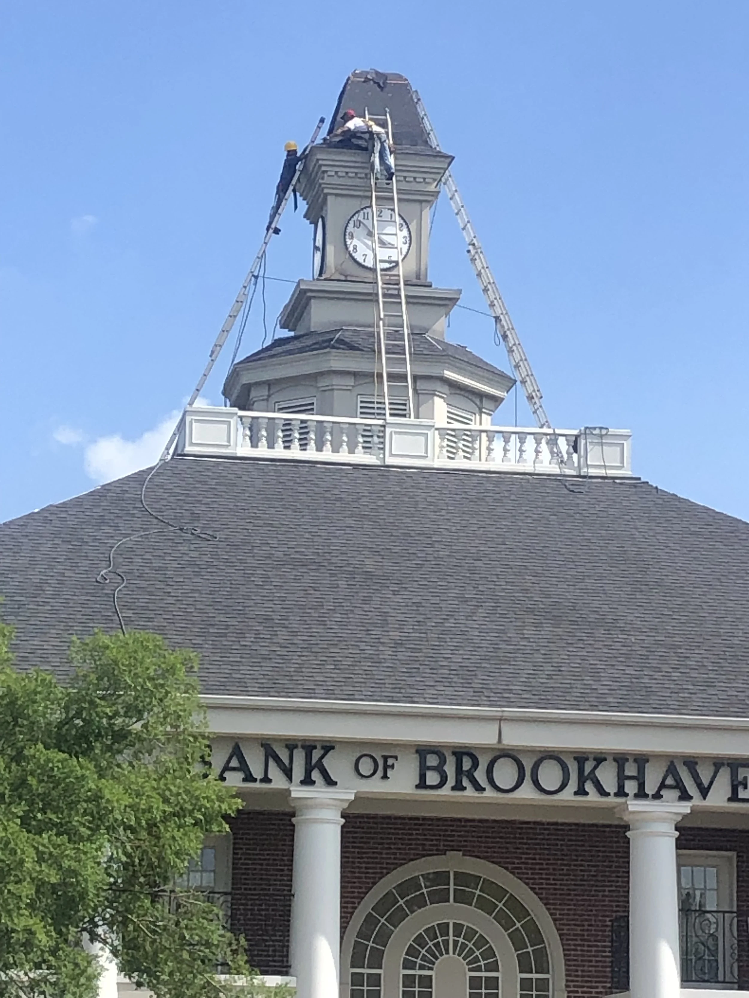 A clock tower under repair at the Bank of Brookhaven, with workers on ladders and a partially covered roof.