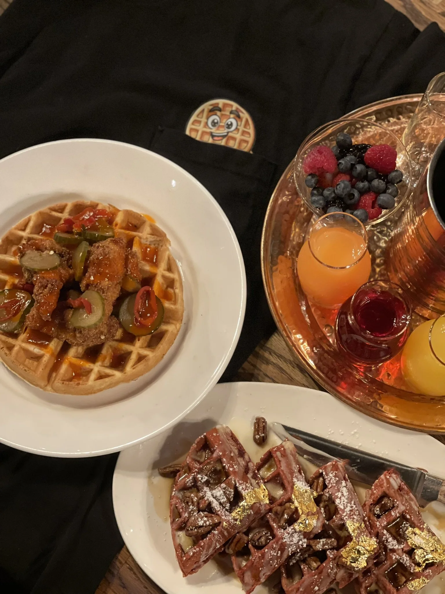A plate of waffles topped with fried chicken and syrup, a plate of waffles with chocolate chips and powdered sugar, and a tray of colorful drinks with berries and orange juice.