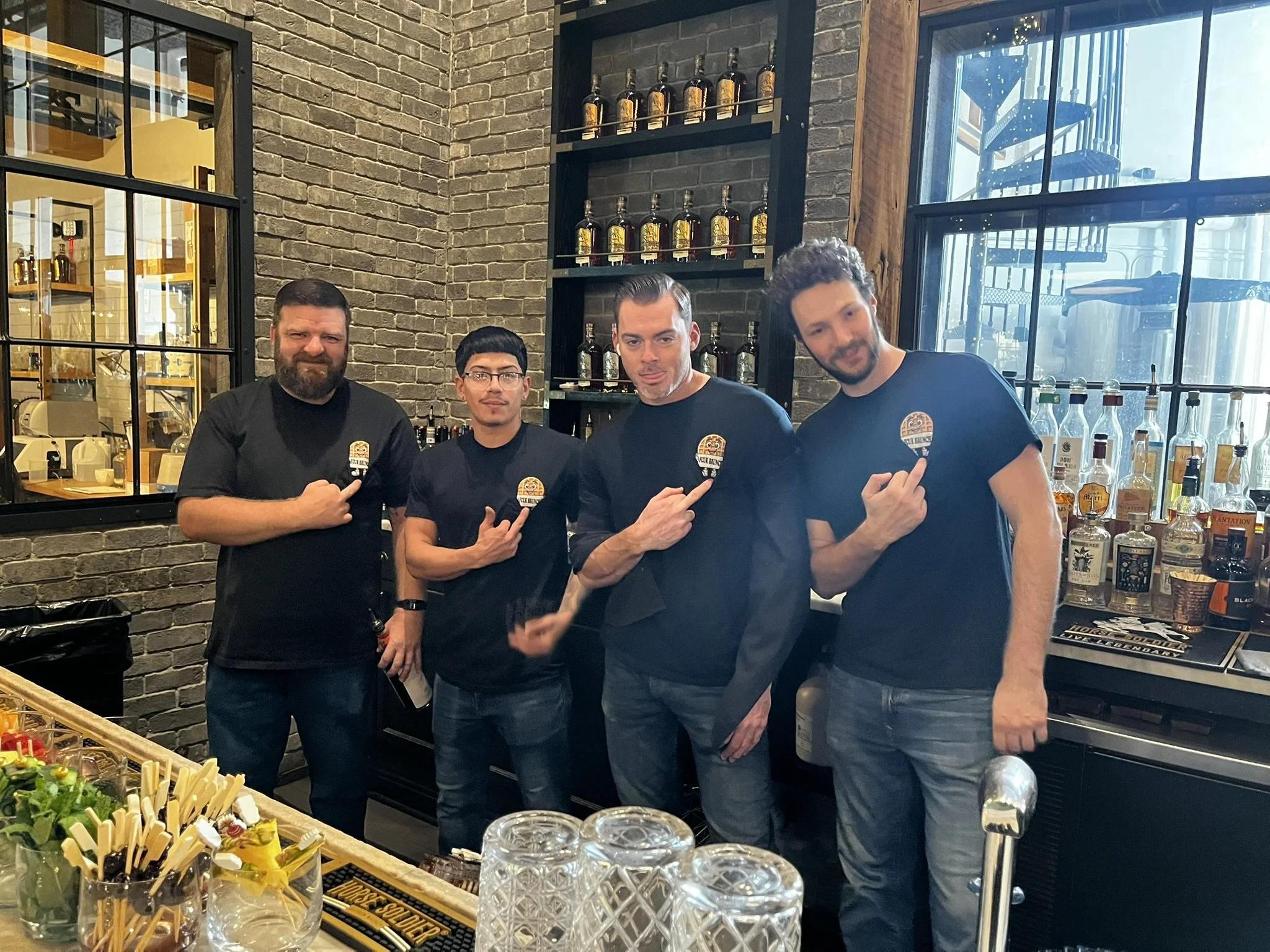 Four men standing behind a bar, all wearing black shirts with a logo. They are pointing to their shirts, and the bar has various bottles of alcohol displayed on shelves behind them. The setting appears to be a brewery or bar with a brick wall and lar