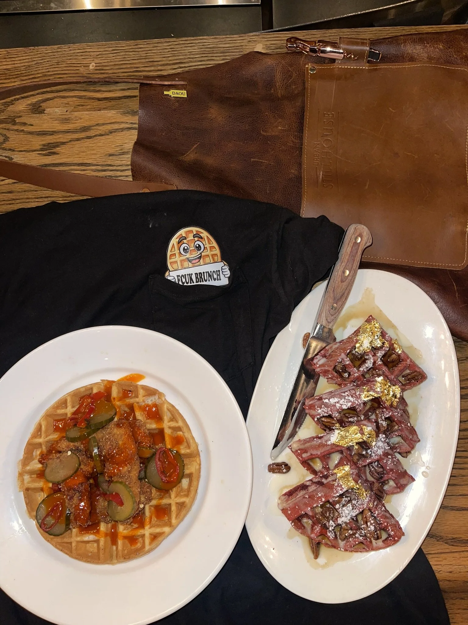 A table with a plate of waffle topped with chili and pickles, and a white platter of sliced cured meat garnished with edible gold leaf, along with a steak knife. A black shirt with an emoji waffle and the text 'FCK BRUNCH' is visible, and a brown lea
