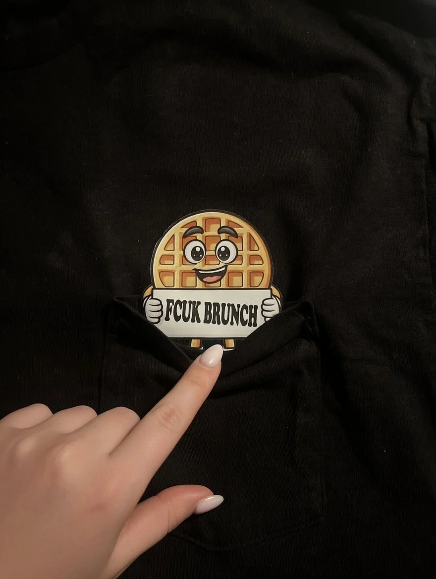 A sticker of a cartoon waffle holding a sign that says 'F*CK BRUNCH' with a person's finger pointing at it.
