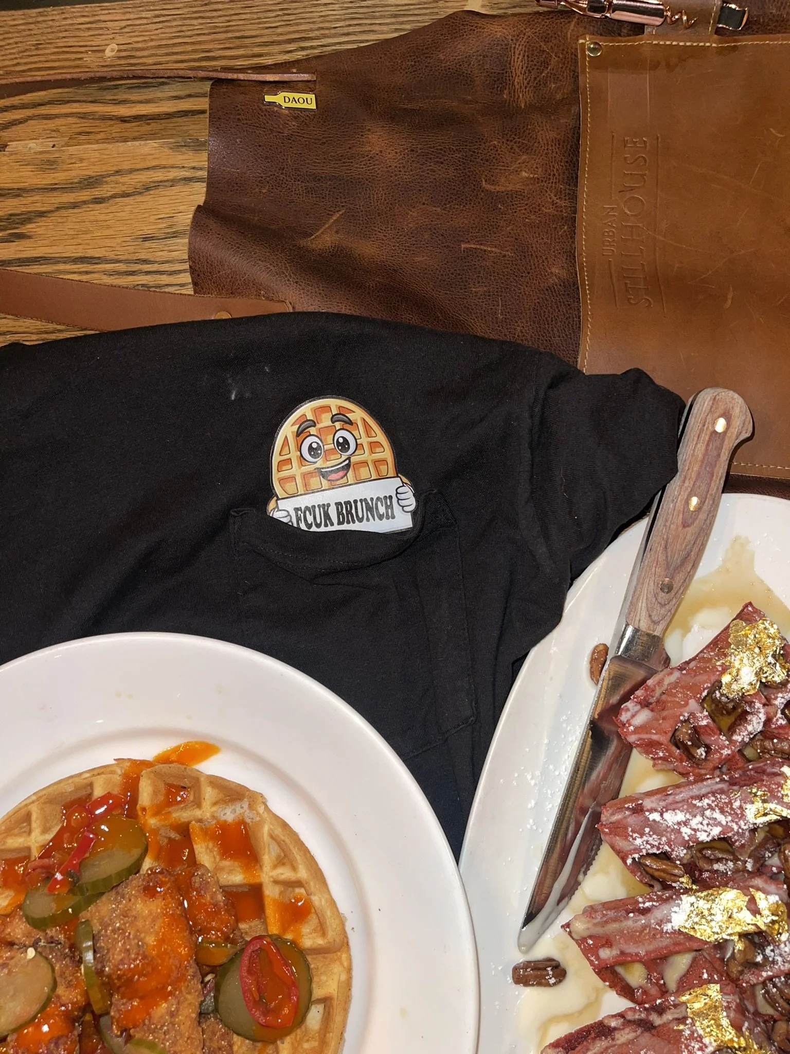 A table with a plate of waffle with fried chicken, pickles, and hot sauce, a charcuterie board with cured meats and gold leaf, a black shirt with a waffle mascot sticker saying 'FCK BRUNCH', a leather bag, and a butter knife.