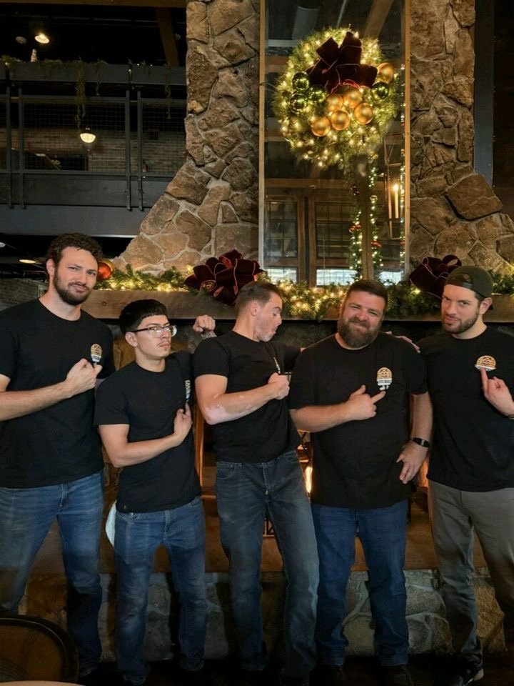 Five men posing in front of a decorated Christmas wreath and window at a restaurant or bar. They are wearing black shirts and making a gesture with their hands over their chests.
