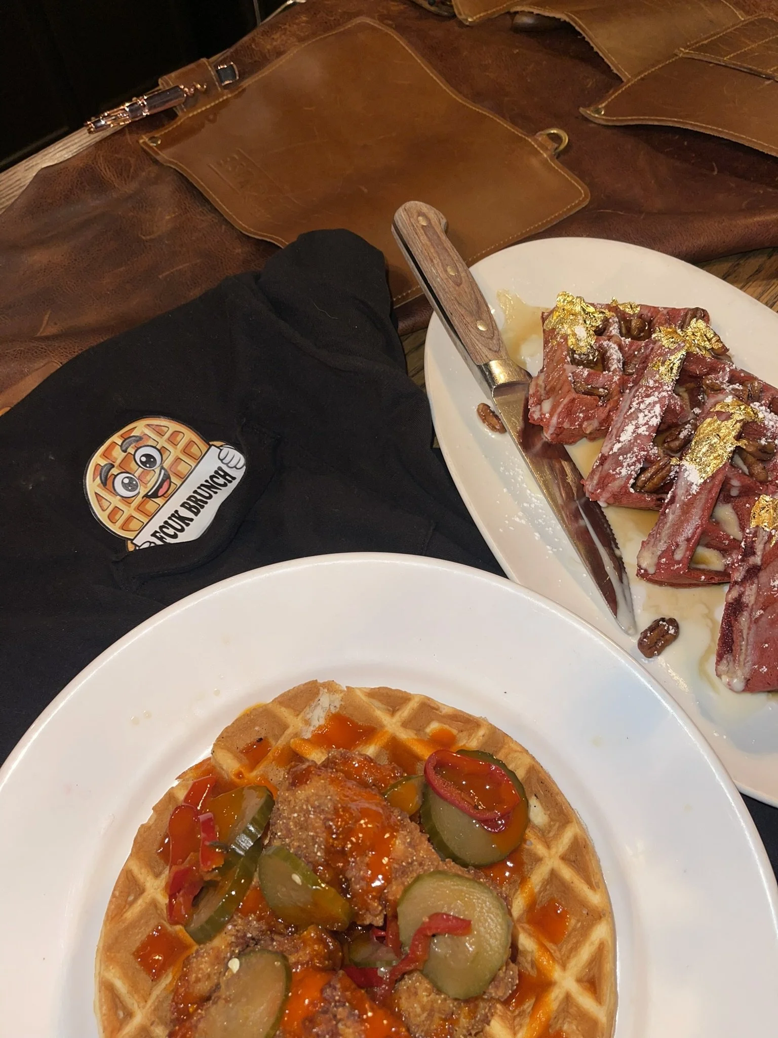A plate with a Belgian waffle topped with fried chicken, pickles, hot sauce, and red peppers. Next to it is a white plate with beef short ribs garnished with gold leaf, pine nuts, and powdered sugar. There's also a brown leather purse and a black pie
