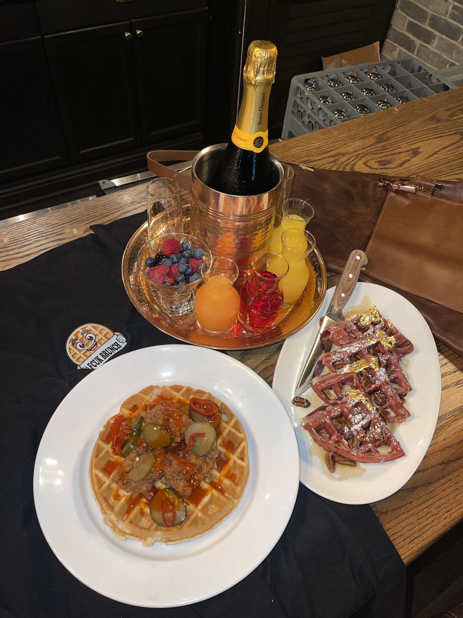 Brunch spread featuring a waffle topped with fruit and syrup, a plate of bacon-wrapped waffles topped with syrup, a tray of assorted drinks, and a bottle of champagne in an ice bucket.
