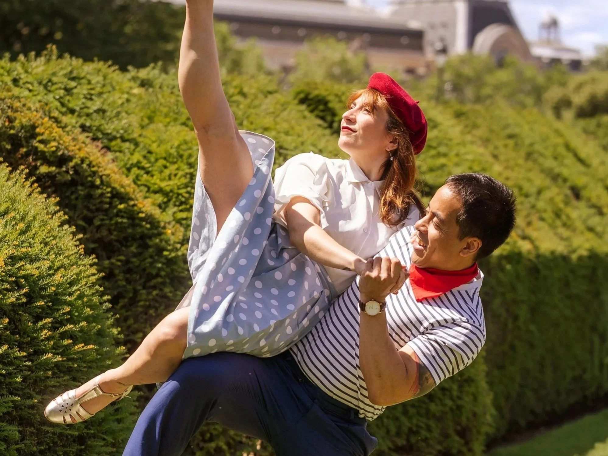 Un homme porte une femme sur ses épaules dans un parc, tous deux souriants, avec un arrière-plan de buissons et un bâtiment à l'horizon.