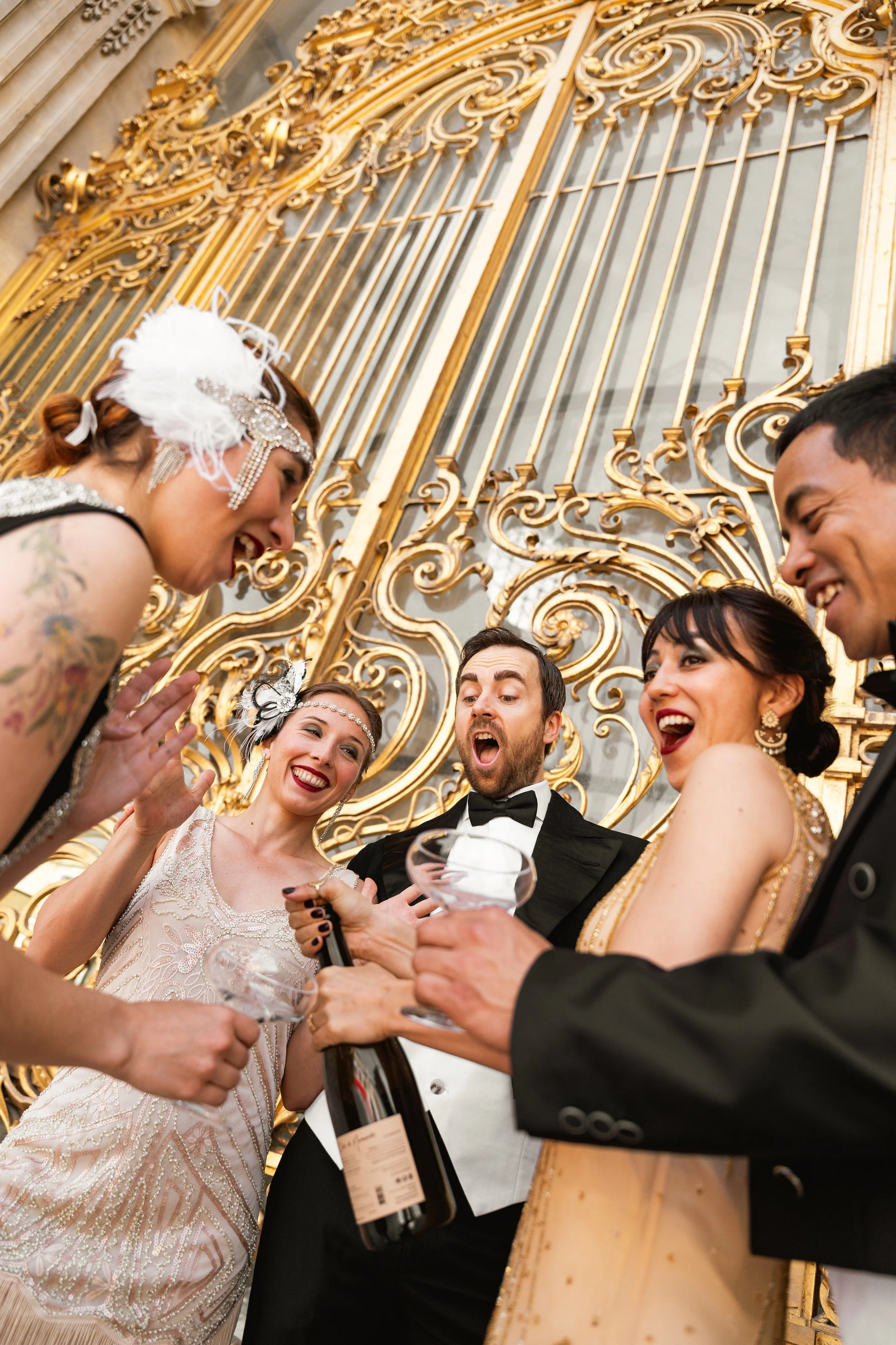 Groupe de personnes habillées à la manière élégante des années 20, célébrant une occasion spéciale, avec des bouteilles de champagne, dans un lieu décoré avec un portail doré ornemental.