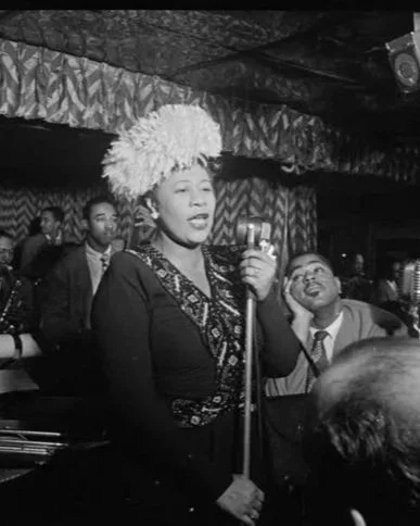 Une femme portant un chapeau de plumes chante ou parle devant un microphone lors d'une performance ou d'une soirée. Autour d'elle, il y a plusieurs personnes, certaines souriantes, dans un cadre intérieur avec des décorations au plafond.