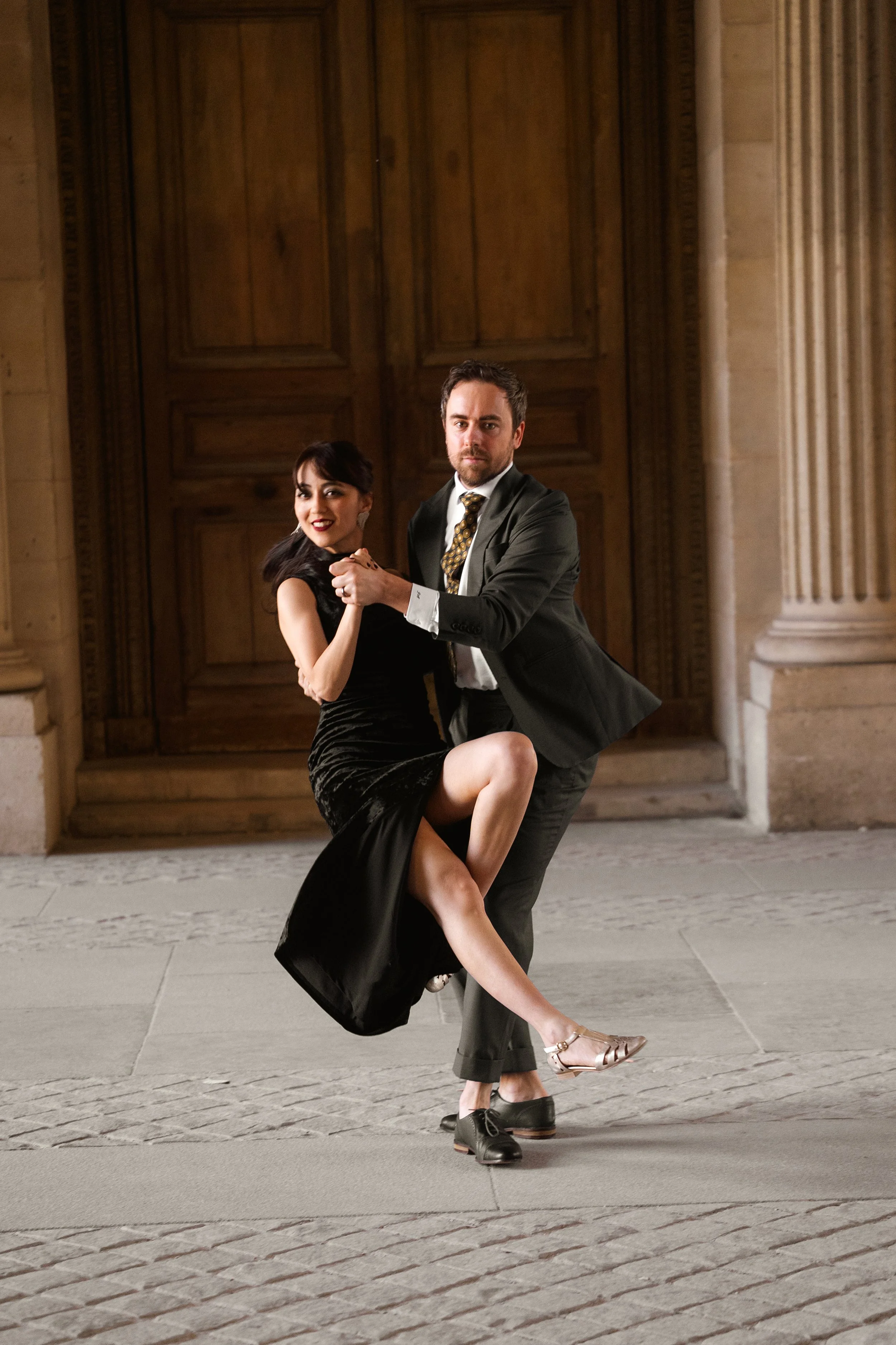 Un homme et une femme dansent ensemble, en tenue élégante, devant un bâtiment en pierre avec de grands portes en bois.