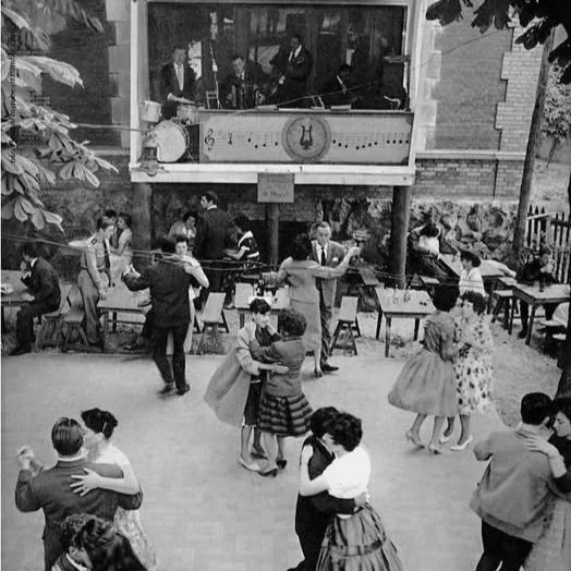 Une scène de danse en plein air avec des couples qui dansent, des musiciens dans une loge supérieure et des spectateurs en arrière-plan, probablement dans les années 1950.
