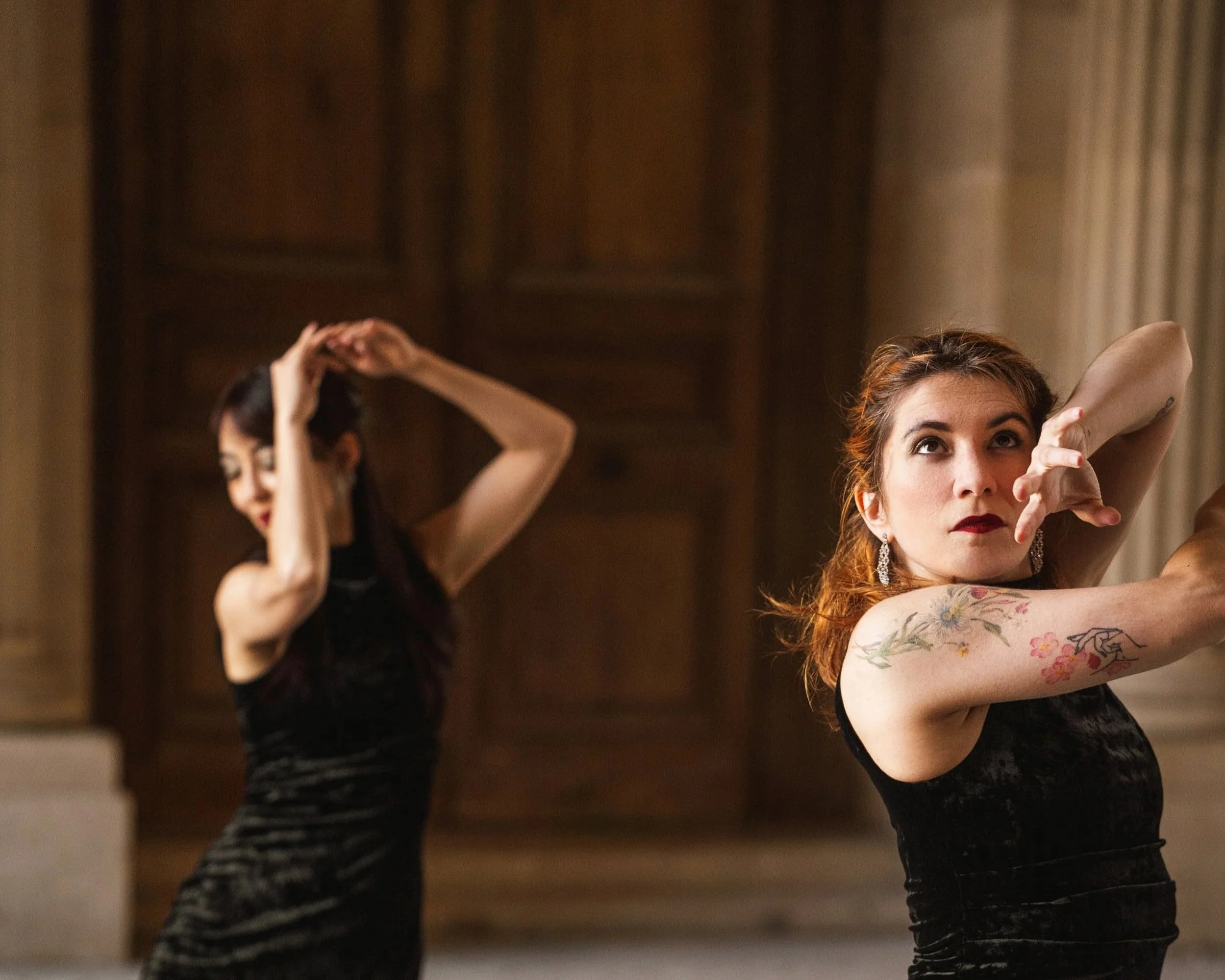 Deux femmes dansent dans une pièce élégante, l'une en avant-plan avec des tatouages sur le bras, l'autre en arrière-plan, toutes deux en position de danse sophistiquée.