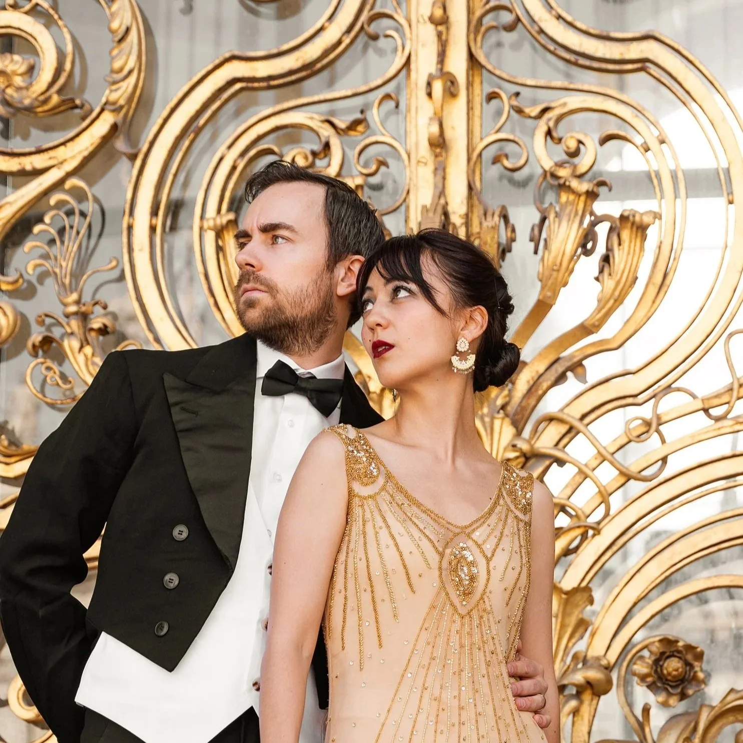 Un homme en smoking noir et une femme en robe dorée élégante posent devant une décoration dorée sophistiquée.