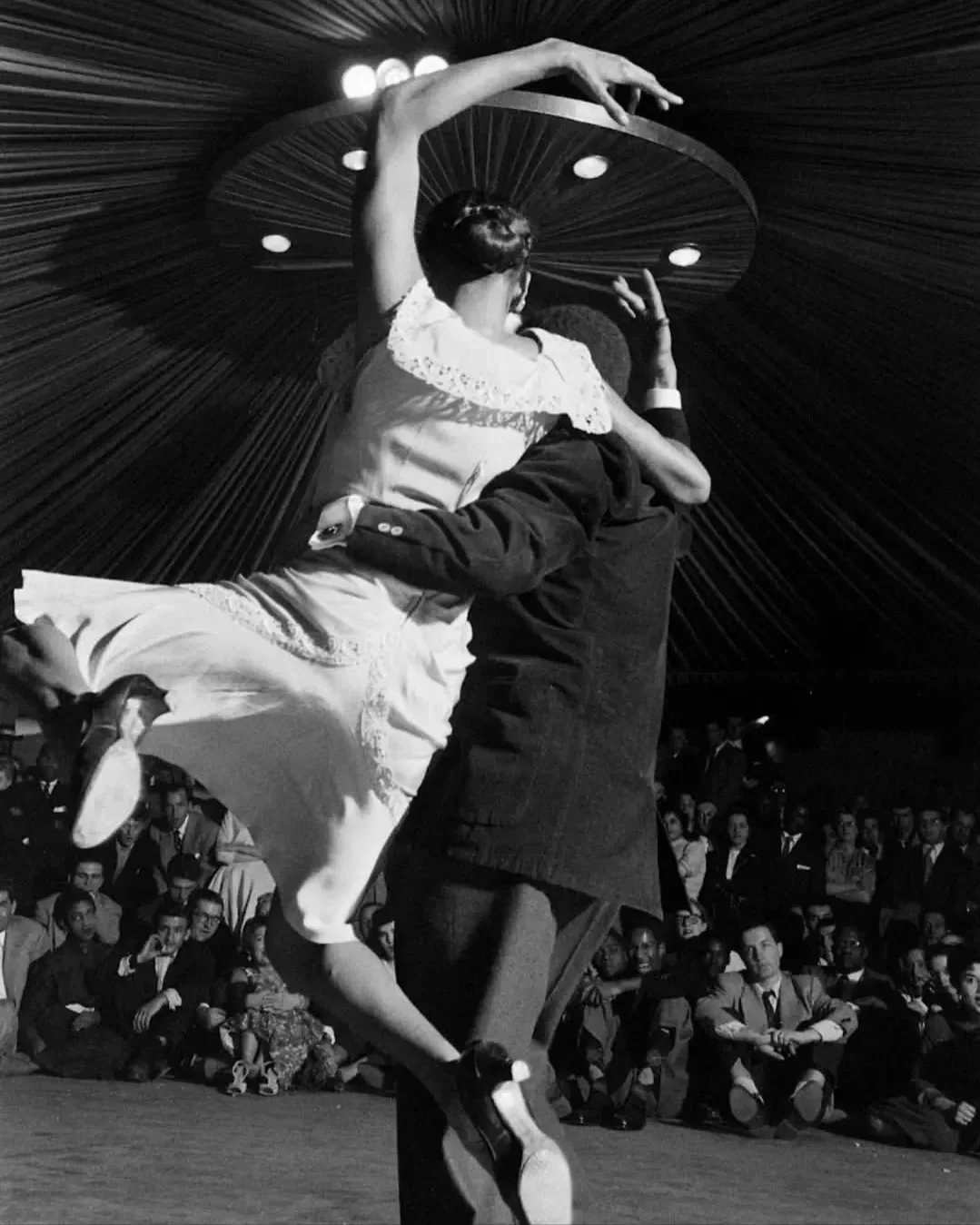 Une femme danse élégamment en duo avec un homme dans une scène de spectacle ou de festival, entourée d'un public assis. La scène est sous un grand chapiteau ou tente.