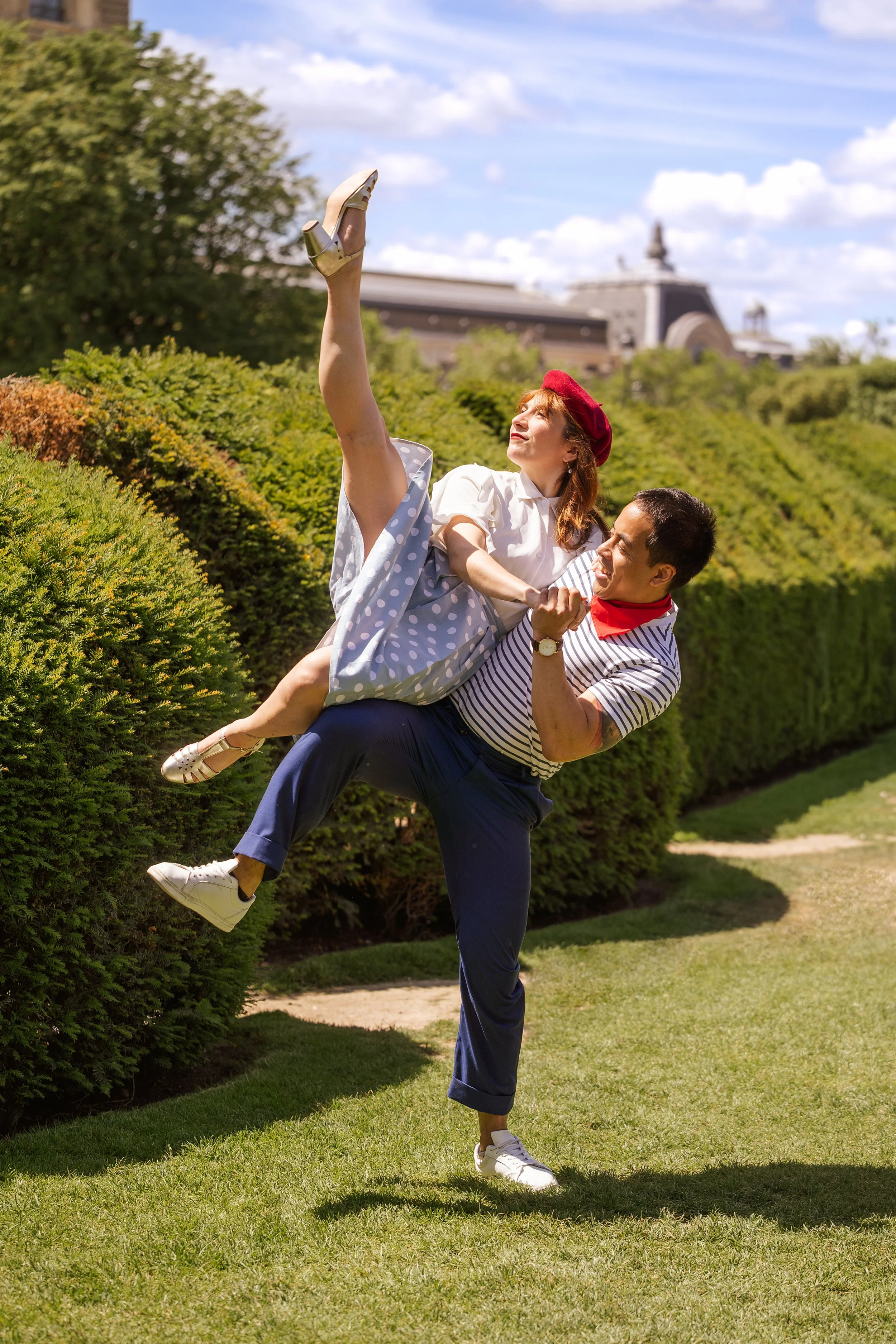 Dans un jardin, un homme vêtu d'une marinère et pantalon bleu et une femme avec une jupe à pois et une beret rouge font un porté de danse swing.