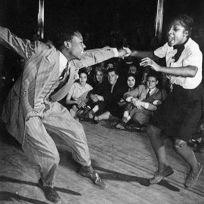 Deux personnes dansent ensemble dans un style swing, avec un groupe de spectateurs en arrière-plan.