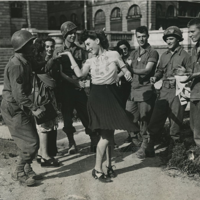 Une femme danse joyeusement entourée de soldats en uniforme, dans une rue en plein jour, semble-t-il pendant la Seconde Guerre mondiale.