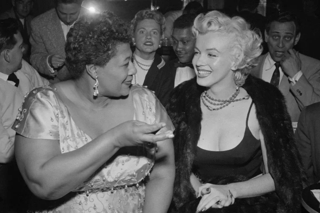 Ella Fitzgerald et Marylin Monroe discutent lors d'une soirée chic, entourées par d'autres personnes en arrière-plan.